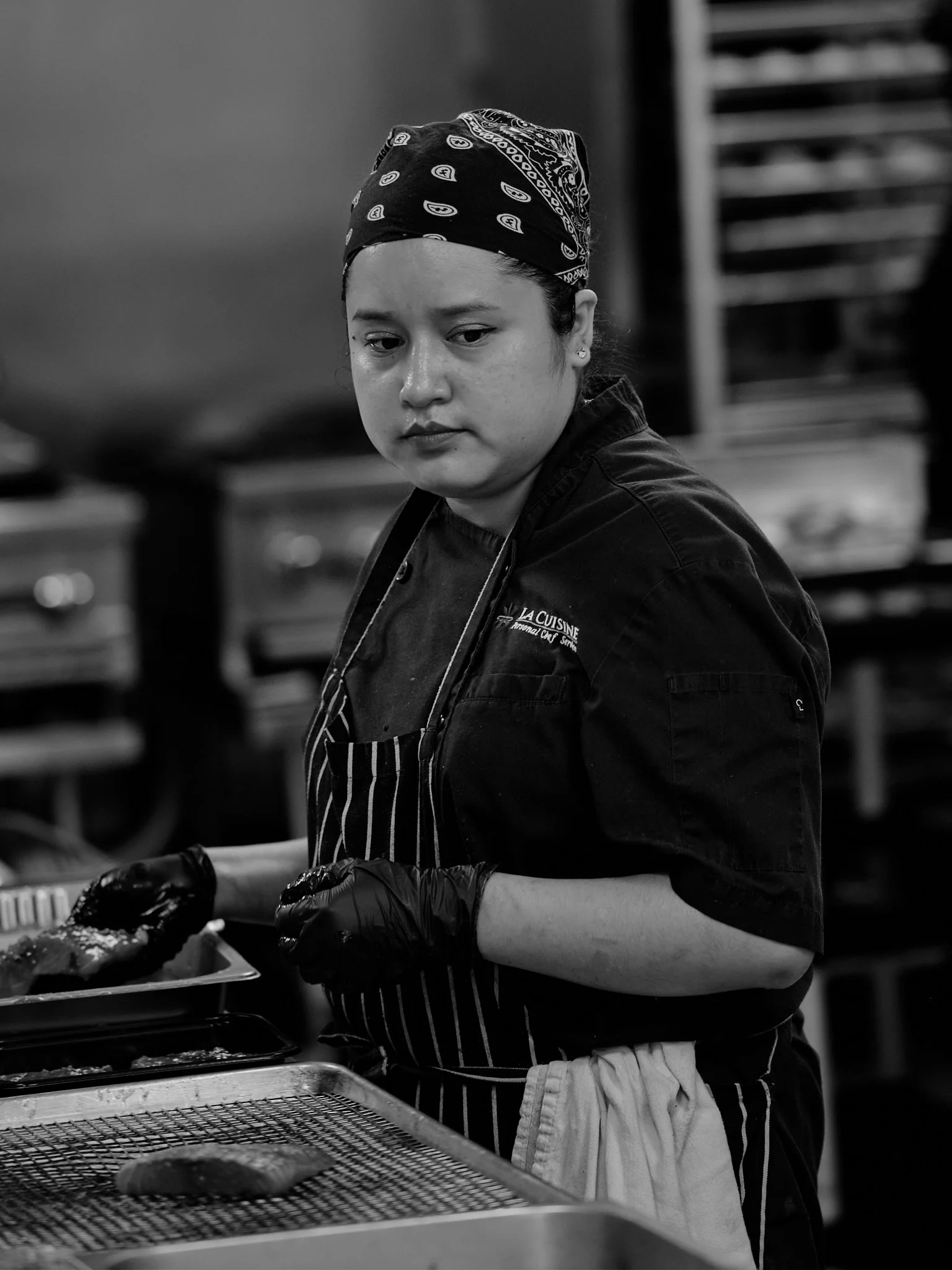 A young woman in a black apron and bandana preparing food in a professional kitchen, with trays of food in front of her.
