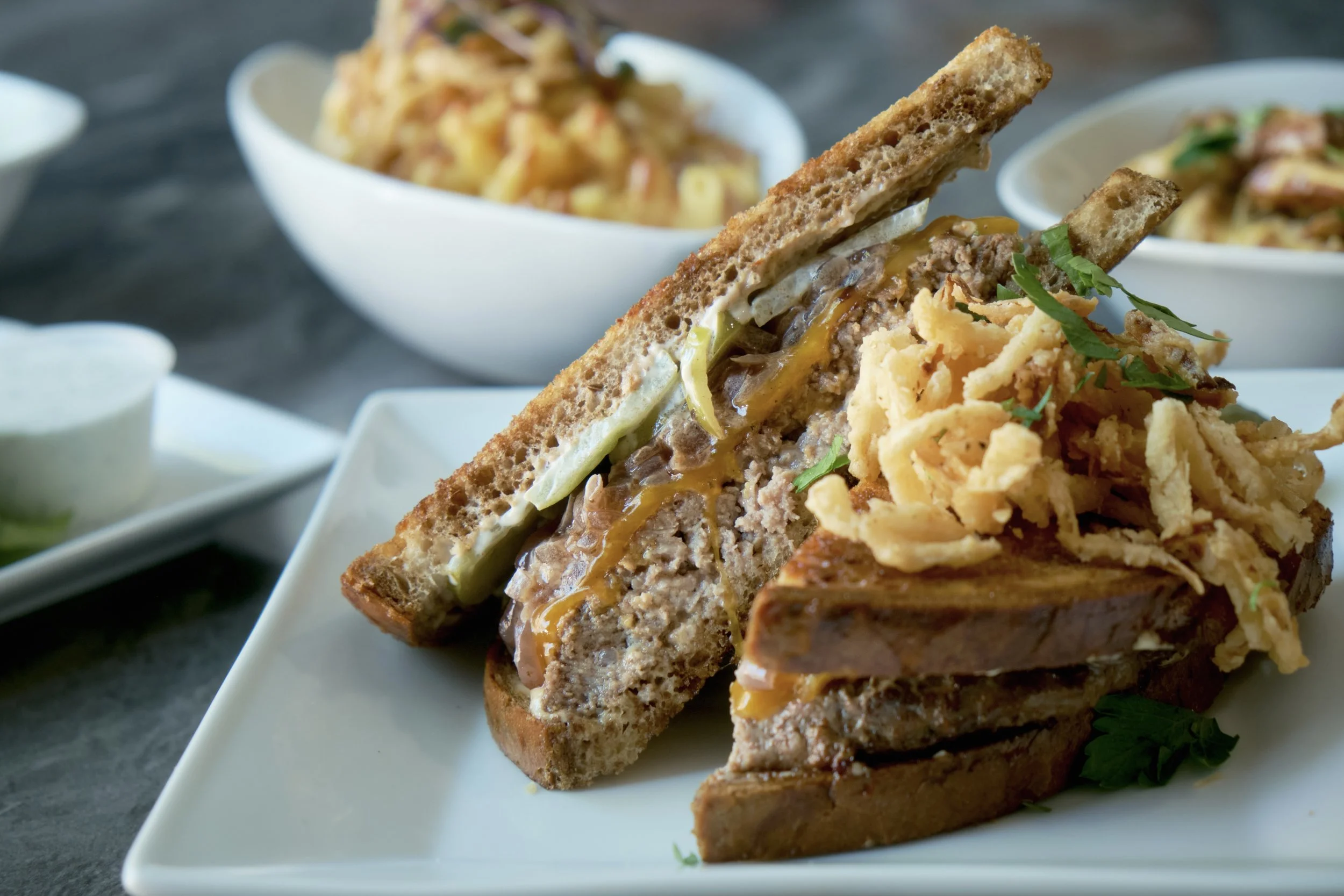 Two slices of a meatloaf sandwich with sauce and crispy onions on top, served on a white plate with a side of shredded lettuce and garnished with parsley.
