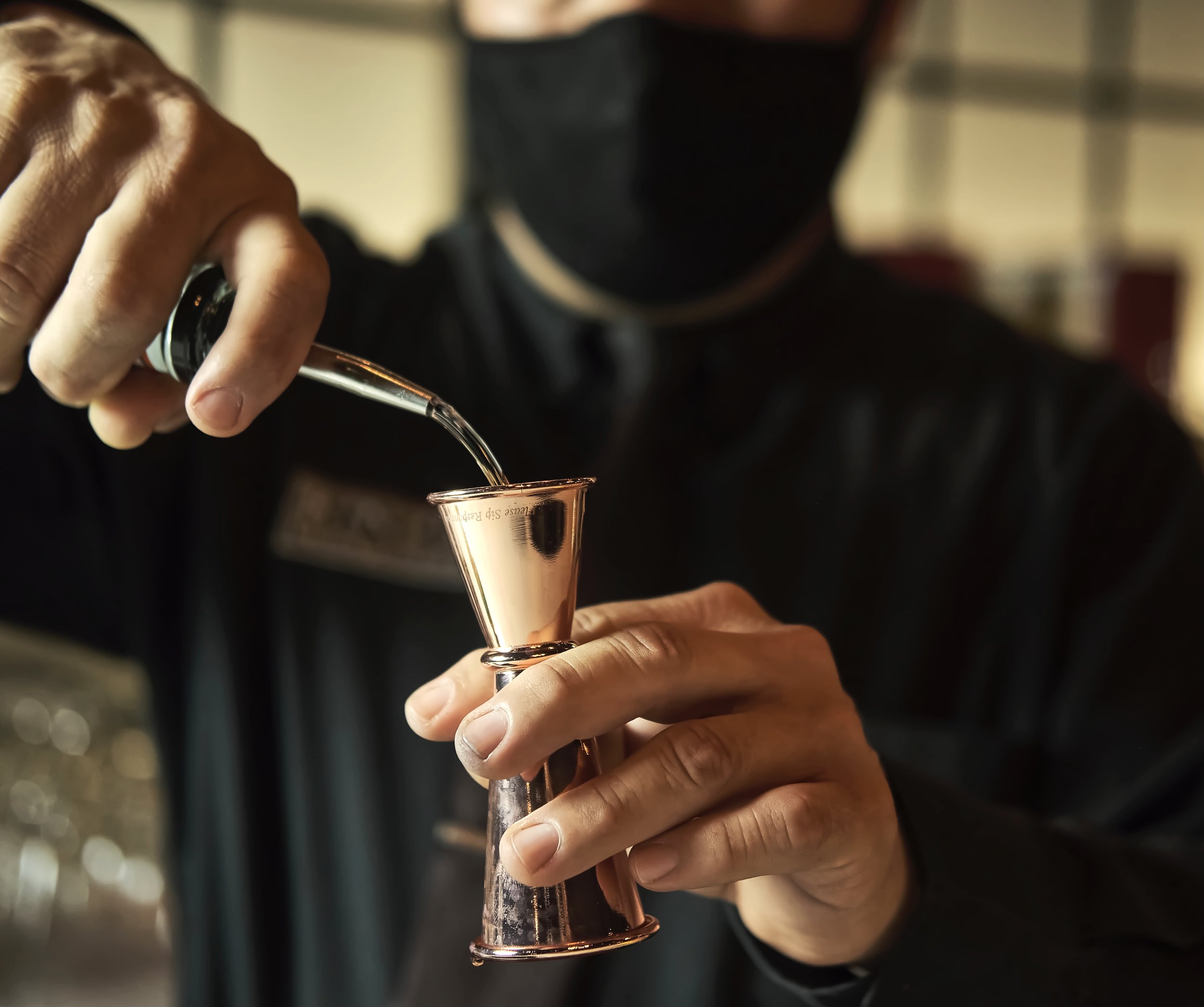 Person pouring a drink into a copper shot glass, wearing a black hoodie and gloves, in a dimly lit setting.