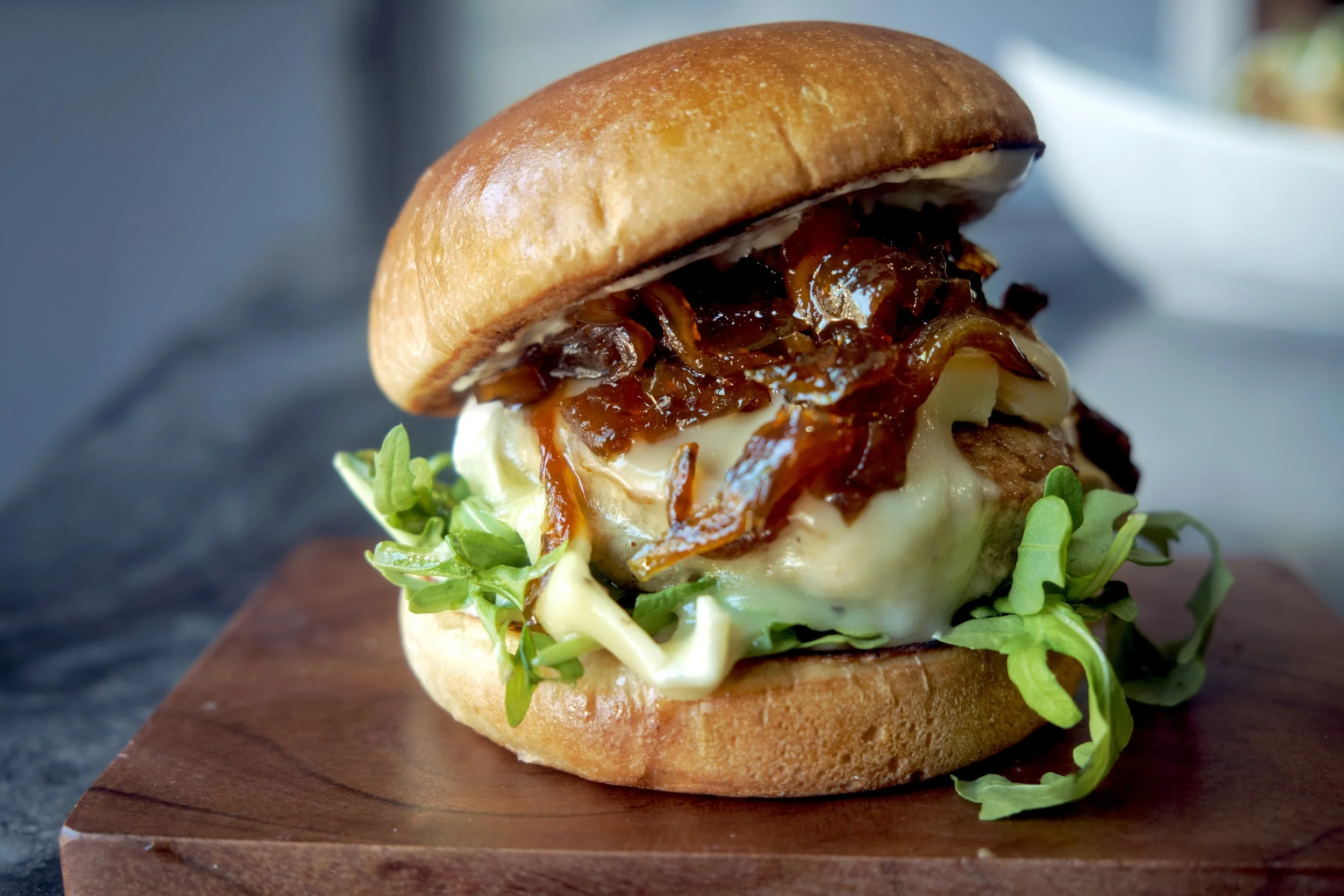 Close-up of a gourmet burger with lettuce, caramelized onions, melted cheese, and barbecue sauce on a toasted bun, served on a wooden cutting board.