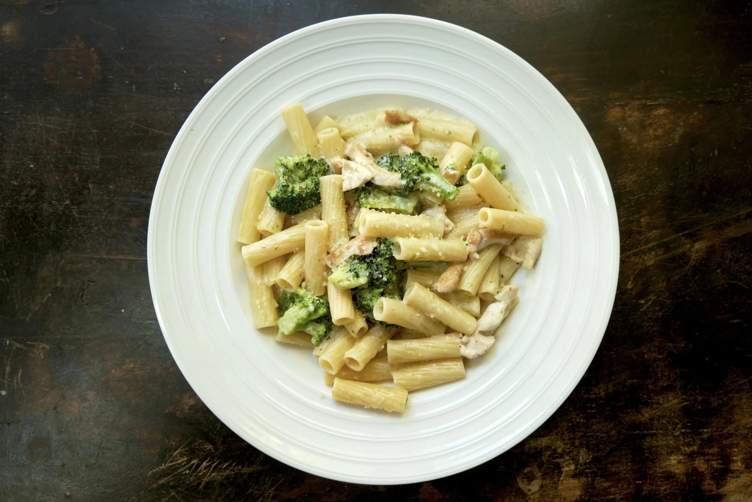 A white plate of cooked pasta with broccoli and chicken on a dark wooden table.