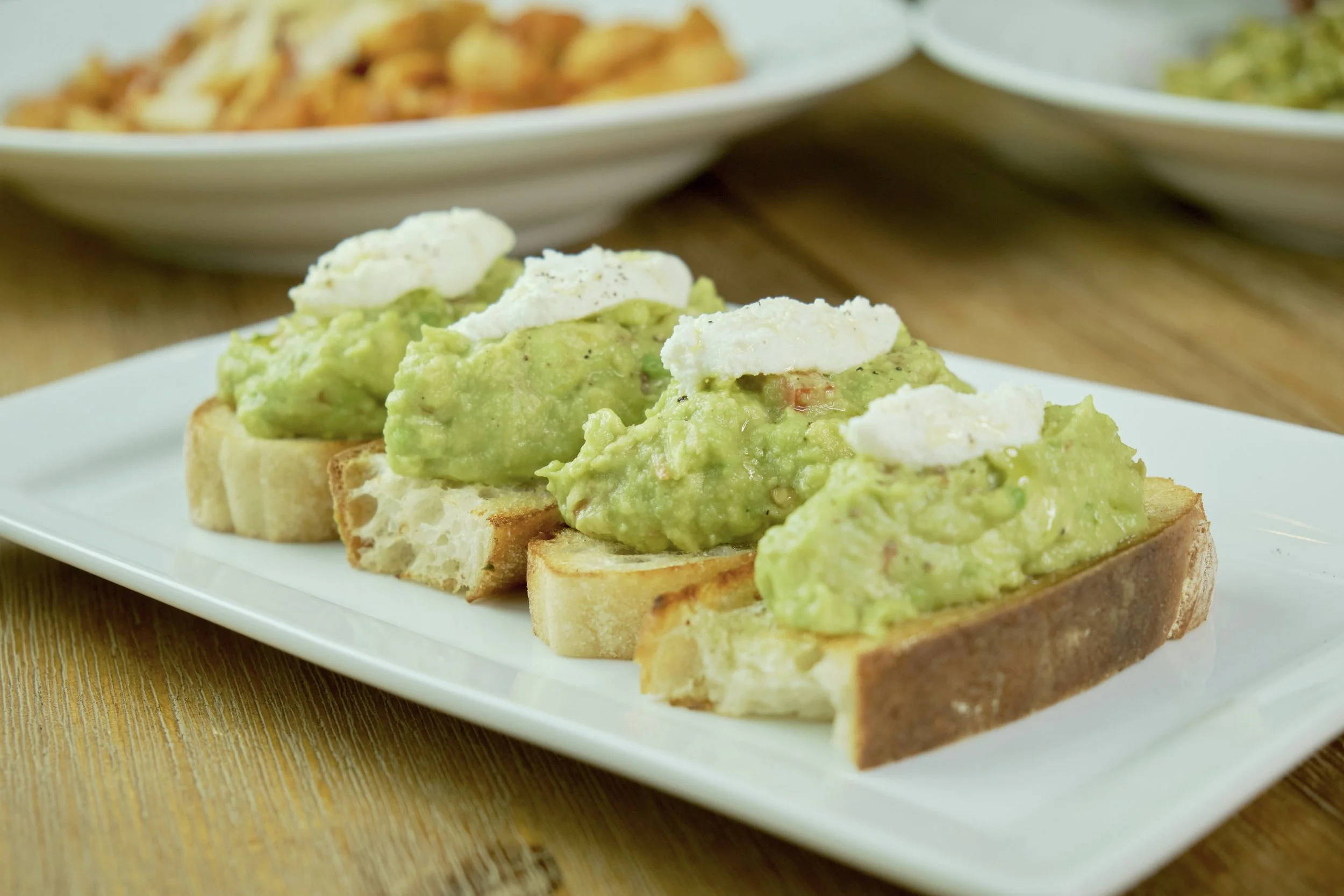 Slices of toasted bread topped with mashed avocado and dollops of sour cream, arranged on a white rectangular plate on a wooden surface.