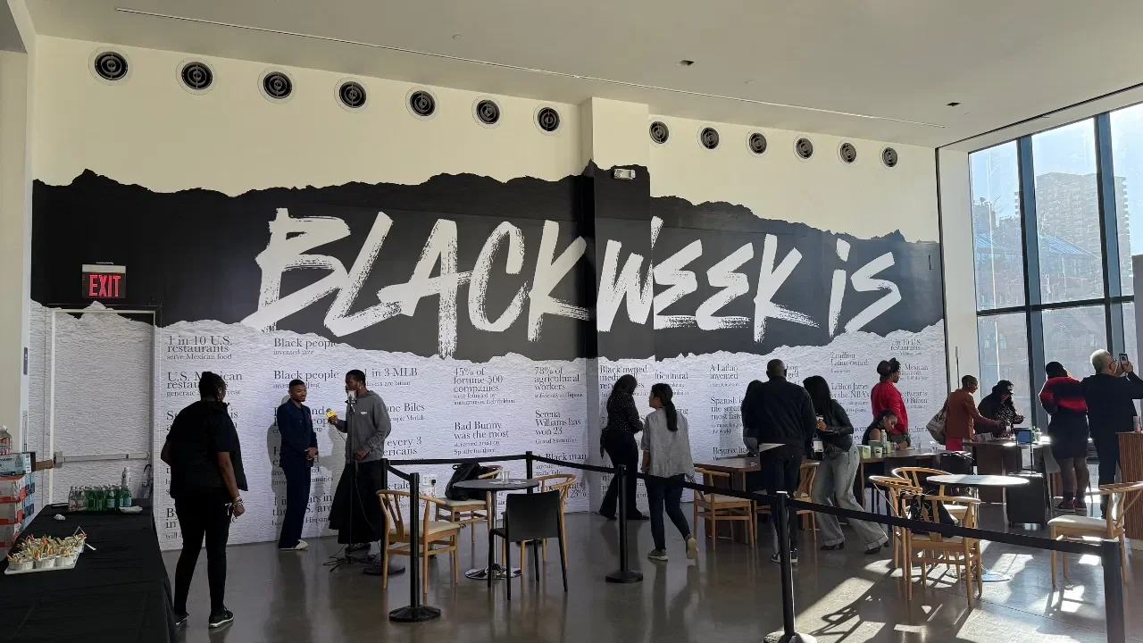 Indoor event space with a large wall mural reading 'Black W  are' in bold white letters on a black background, with people standing and talking near tables and drinks, and large windows on the right side showing a cityscape.