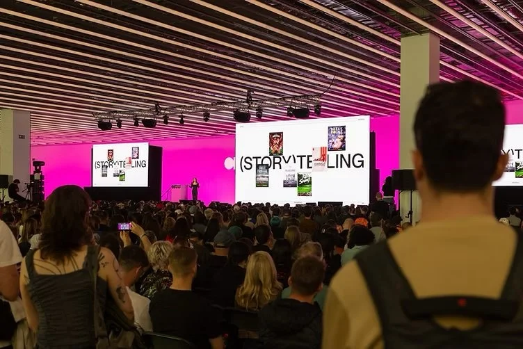 A large audience attending a presentation or conference in a spacious hall with a high ceiling. The stage has two large screens displaying the word "STORYTELLING" with book covers surrounding it, and a speaker standing at a podium.