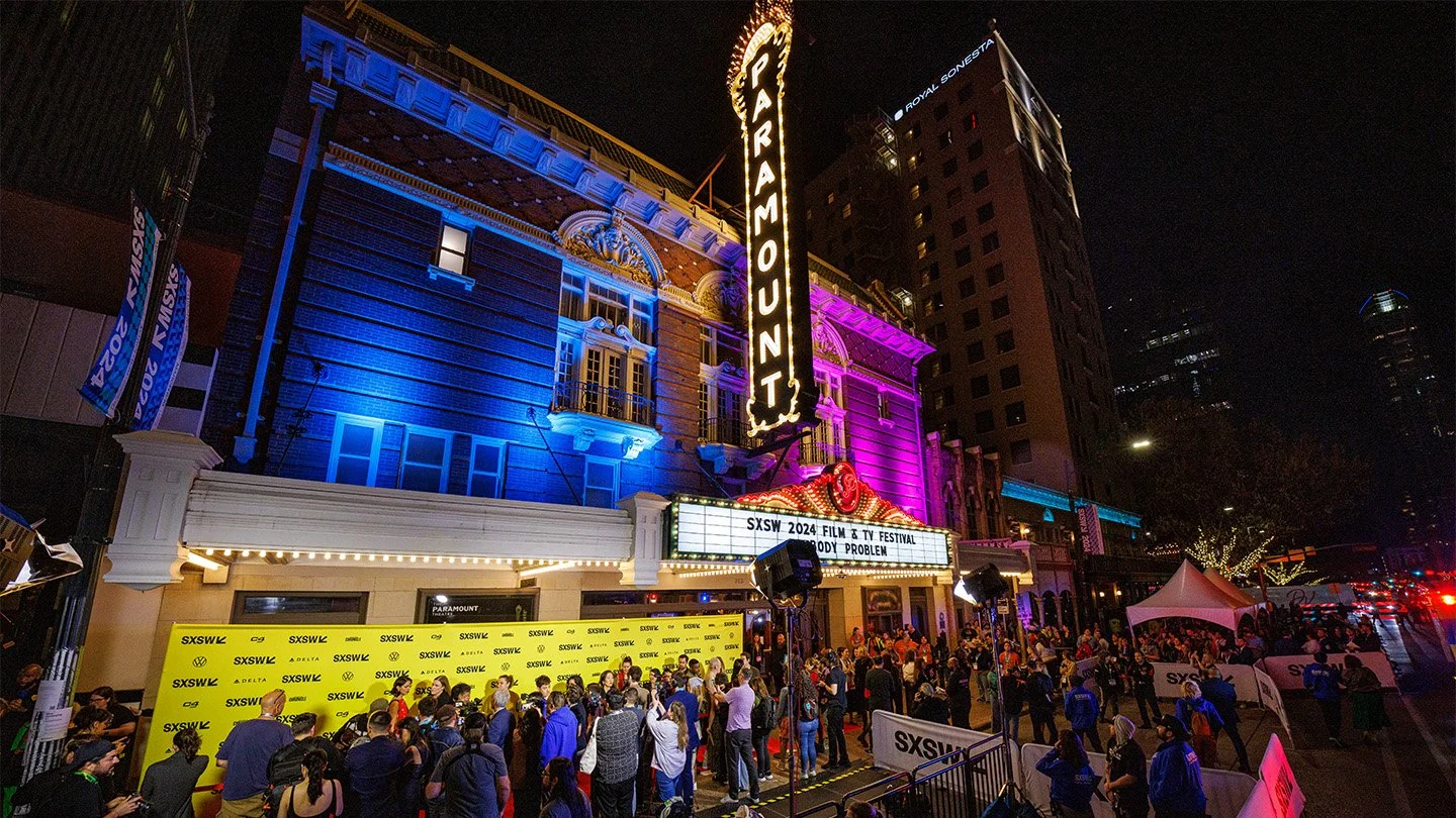 A large crowd of people gathered outside the Paramount theater at SXSW 2024. The theater is lit with colorful neon lights, and a marquee displays the event information. The scene is lively with various tents and signage along the street at night.