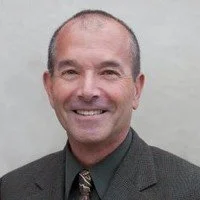 A smiling middle-aged man wearing a suit and tie, standing against a plain background.