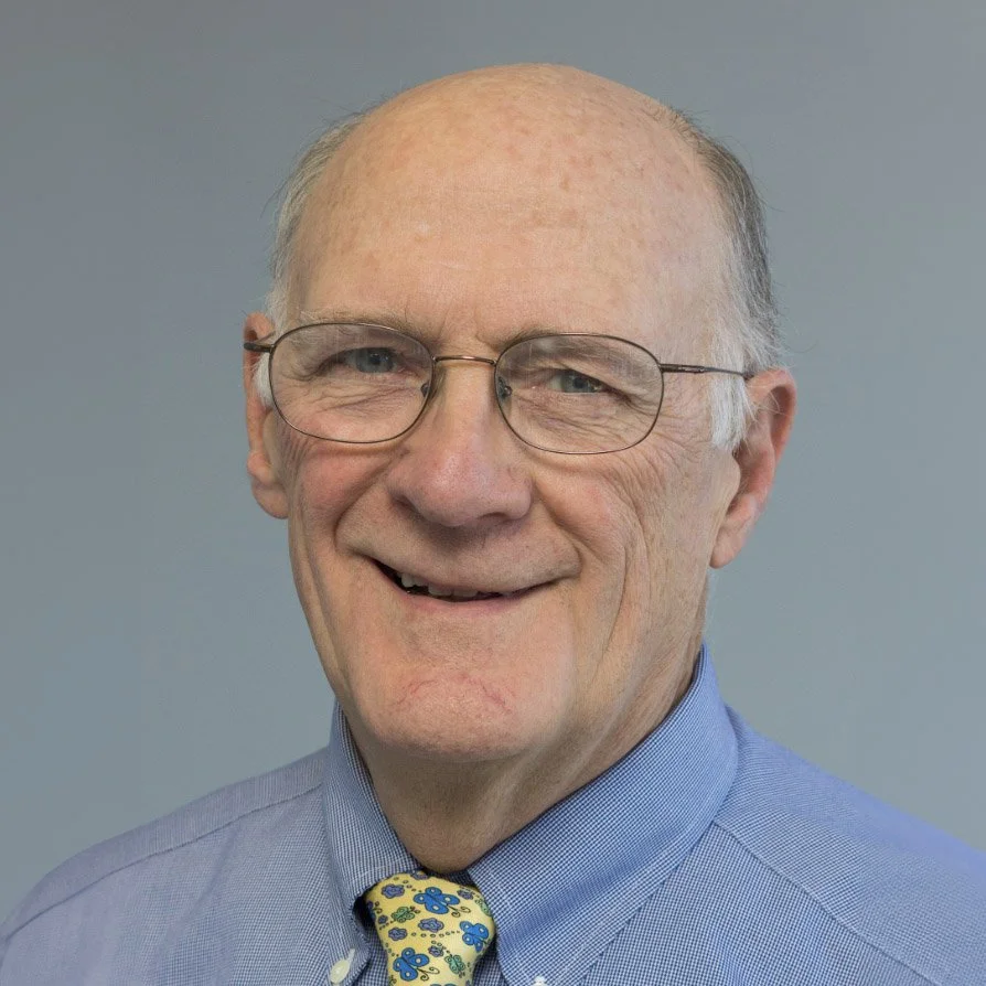 A middle-aged man with glasses, wearing a light blue dress shirt and a yellow tie with blue flowers, smiling against a plain gray background.