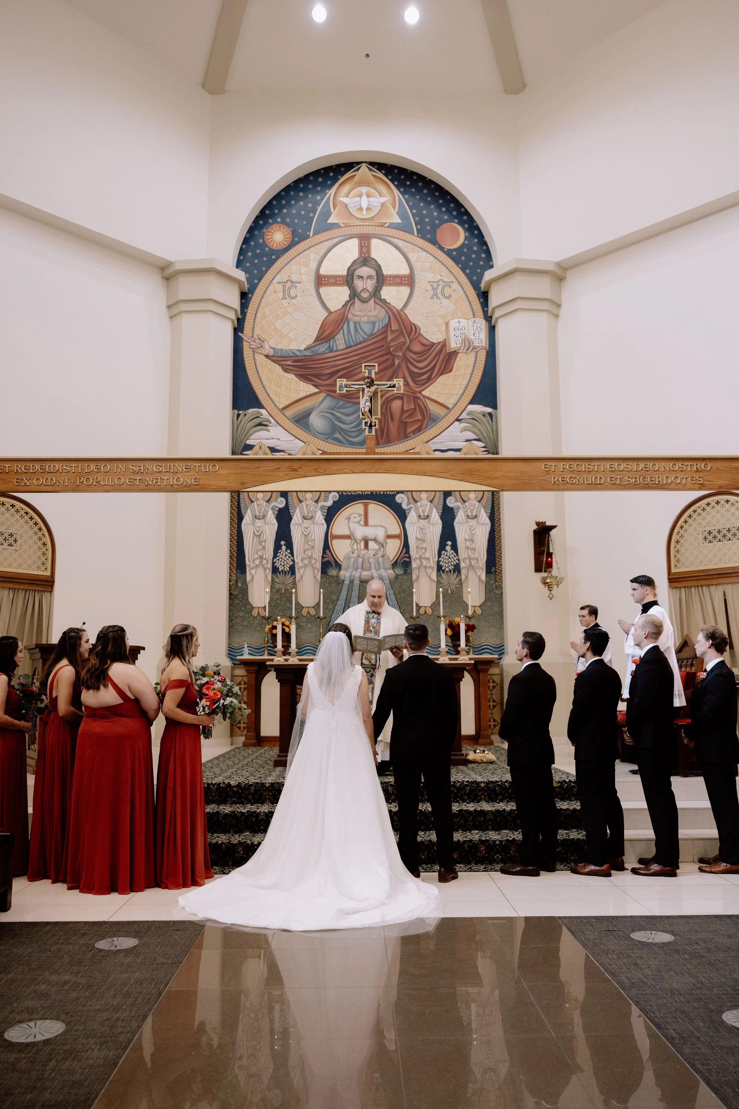 A wedding ceremony taking place inside a church, with a bride and groom standing at the altar facing a priest. Bridesmaids in red dresses and groomsmen in black suits stand on either side of the couple. The church's background features a large religious mural of Jesus Christ and angels, with a wooden cross and lit candles on the altar.