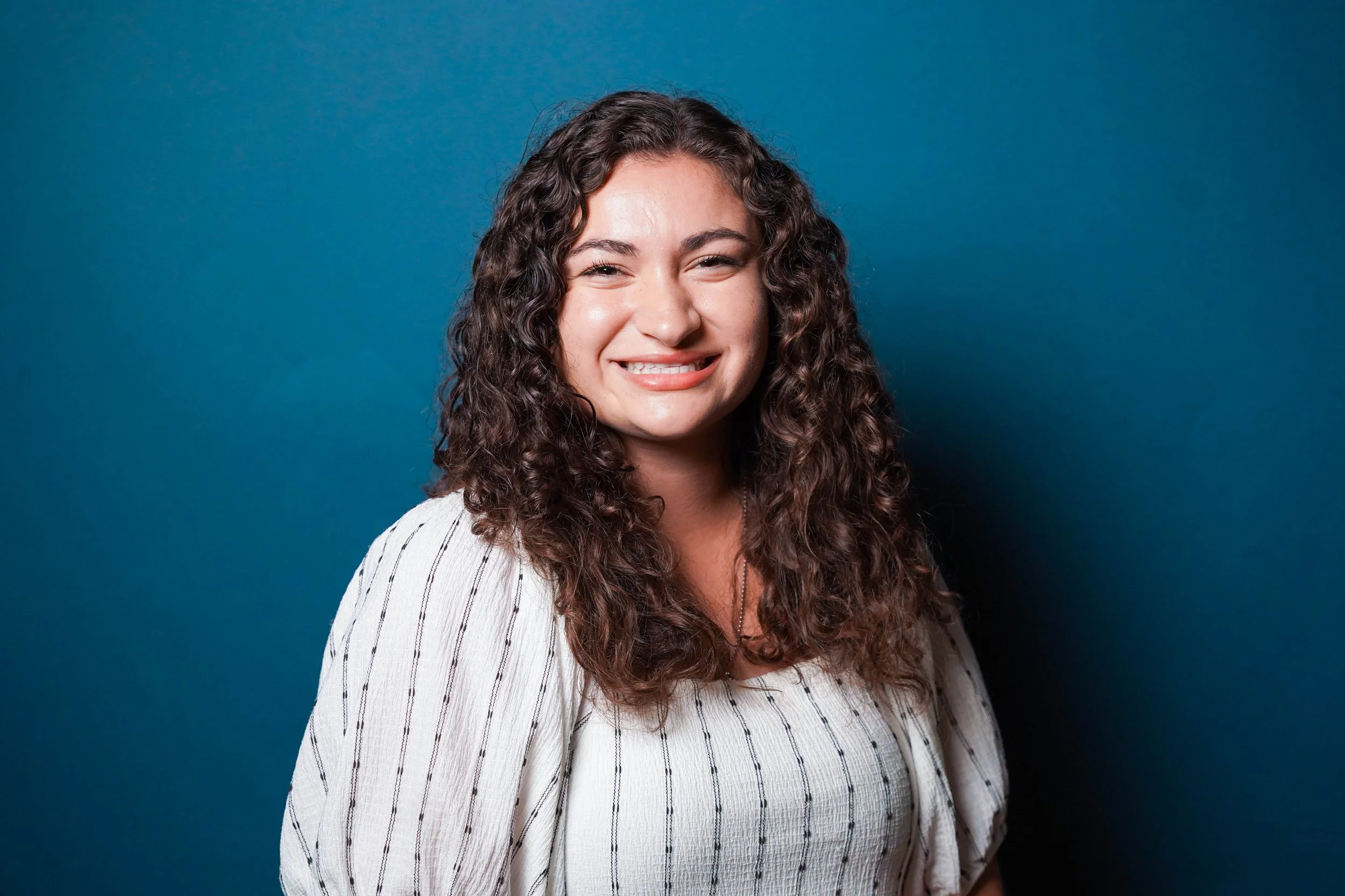 A young woman with long, curly brown hair smiling against a blue background.