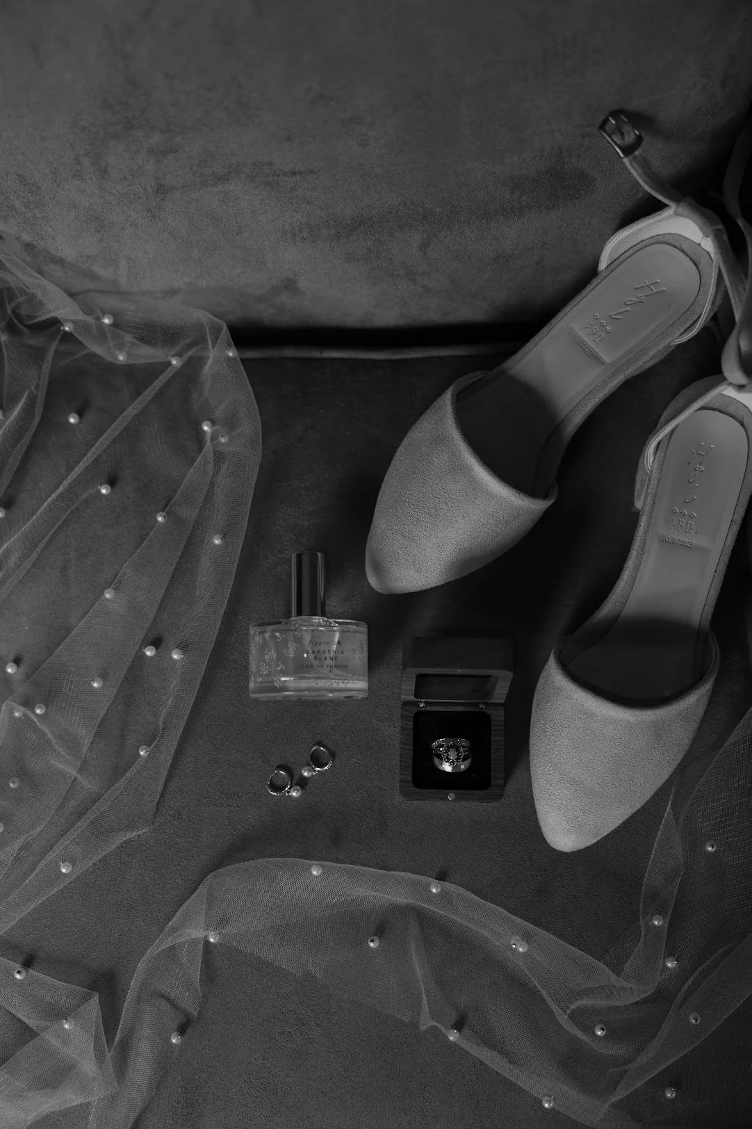 Gray high-heeled shoes, a jar of perfume, rings, and pearls on a soft surface with a sheer, pearl-embellished fabric.