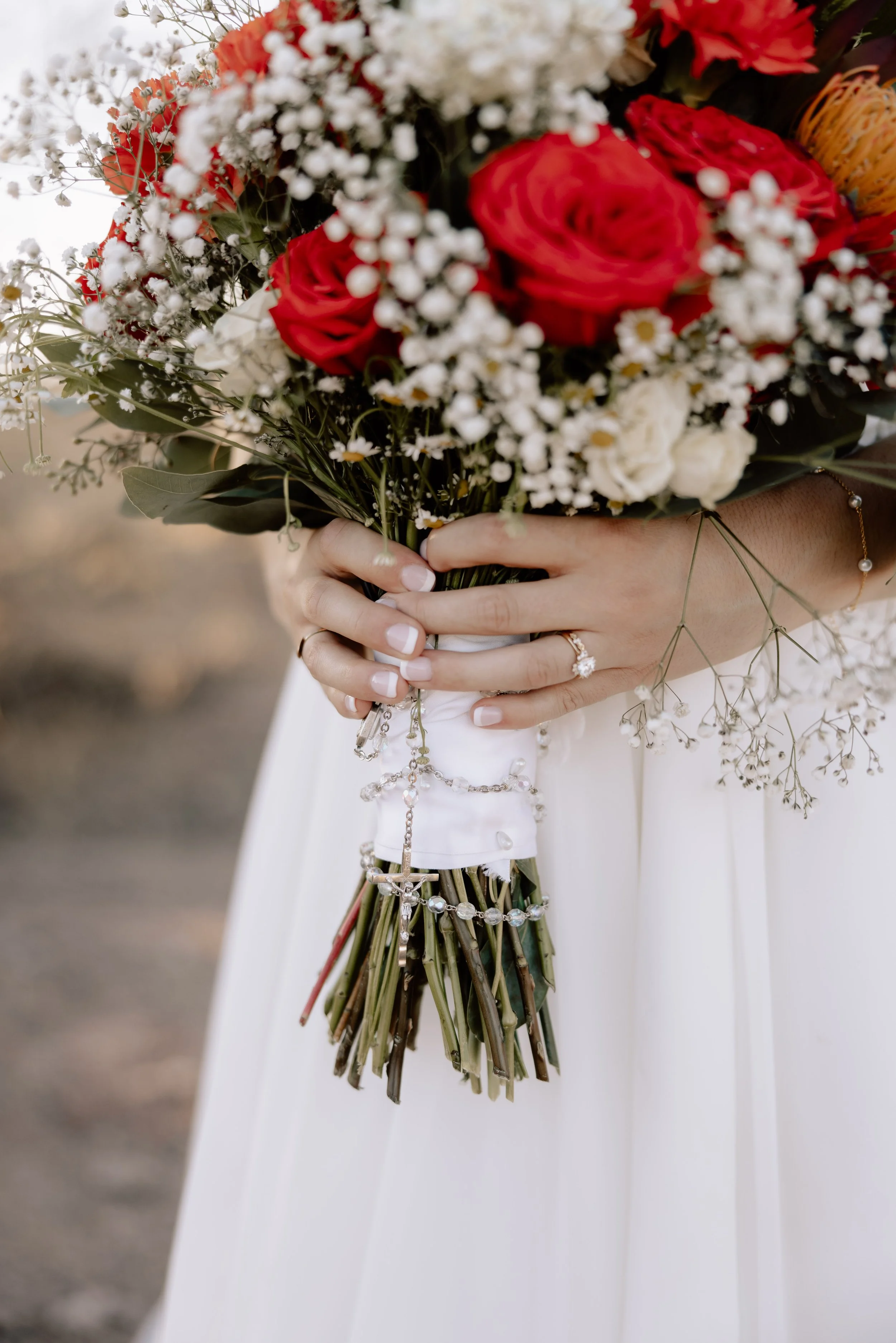 Close-up of a person's hands holding a bridal bouquet with red roses, white flowers, and baby's breath, wrapped in white fabric with jewelry, against a blurred outdoor background.