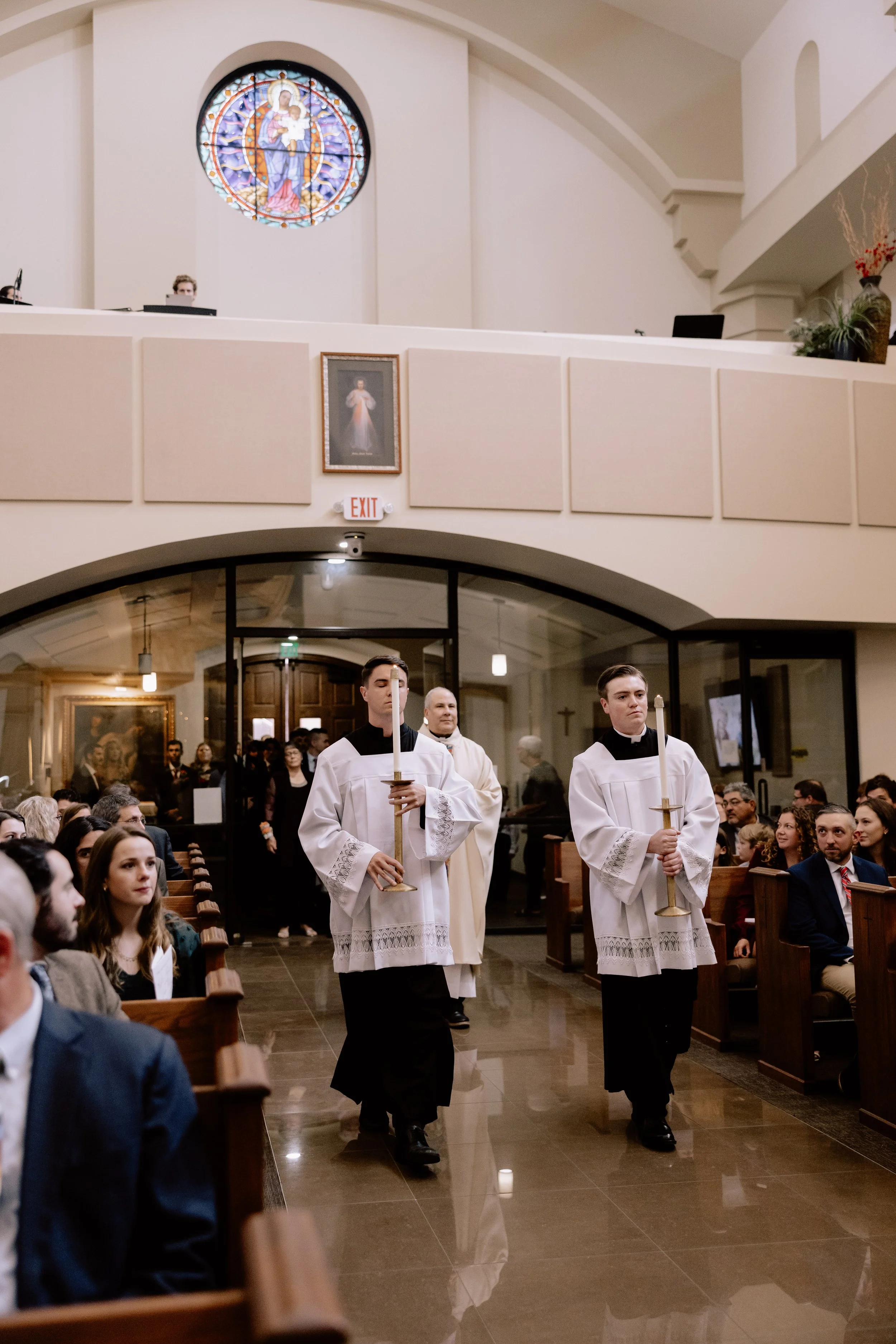 Two altar servers in white robes with black collars holding candles and a cross walk down the aisle of a church during a religious ceremony. The congregation is seated on wooden pews, and a priest stands behind them. The church has a stained glass window of a religious figure and a framed picture of Jesus Christ on the wall.