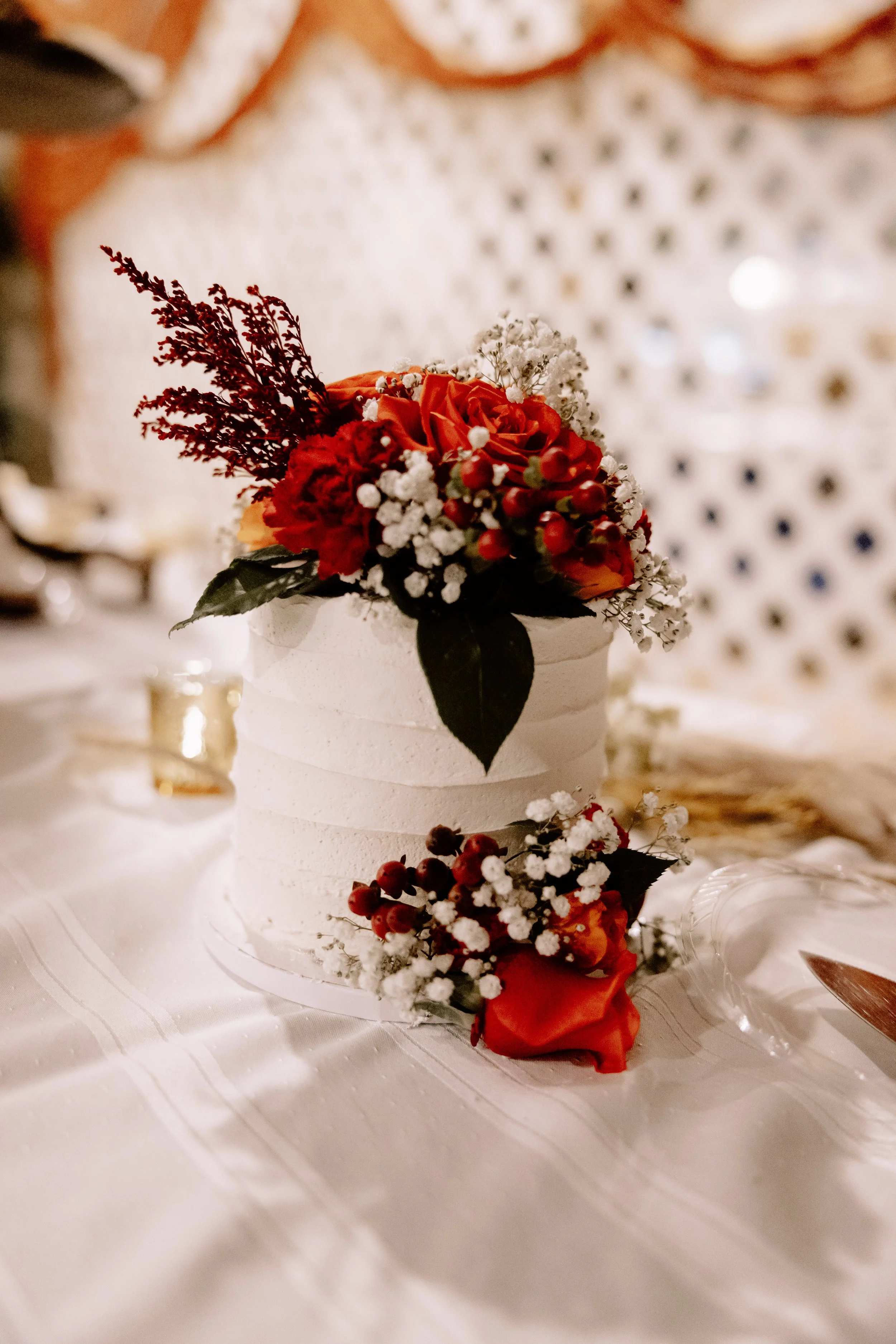A white frosted cake decorated with red and white flowers, dark leaves, and berries, placed on a white tablecloth.