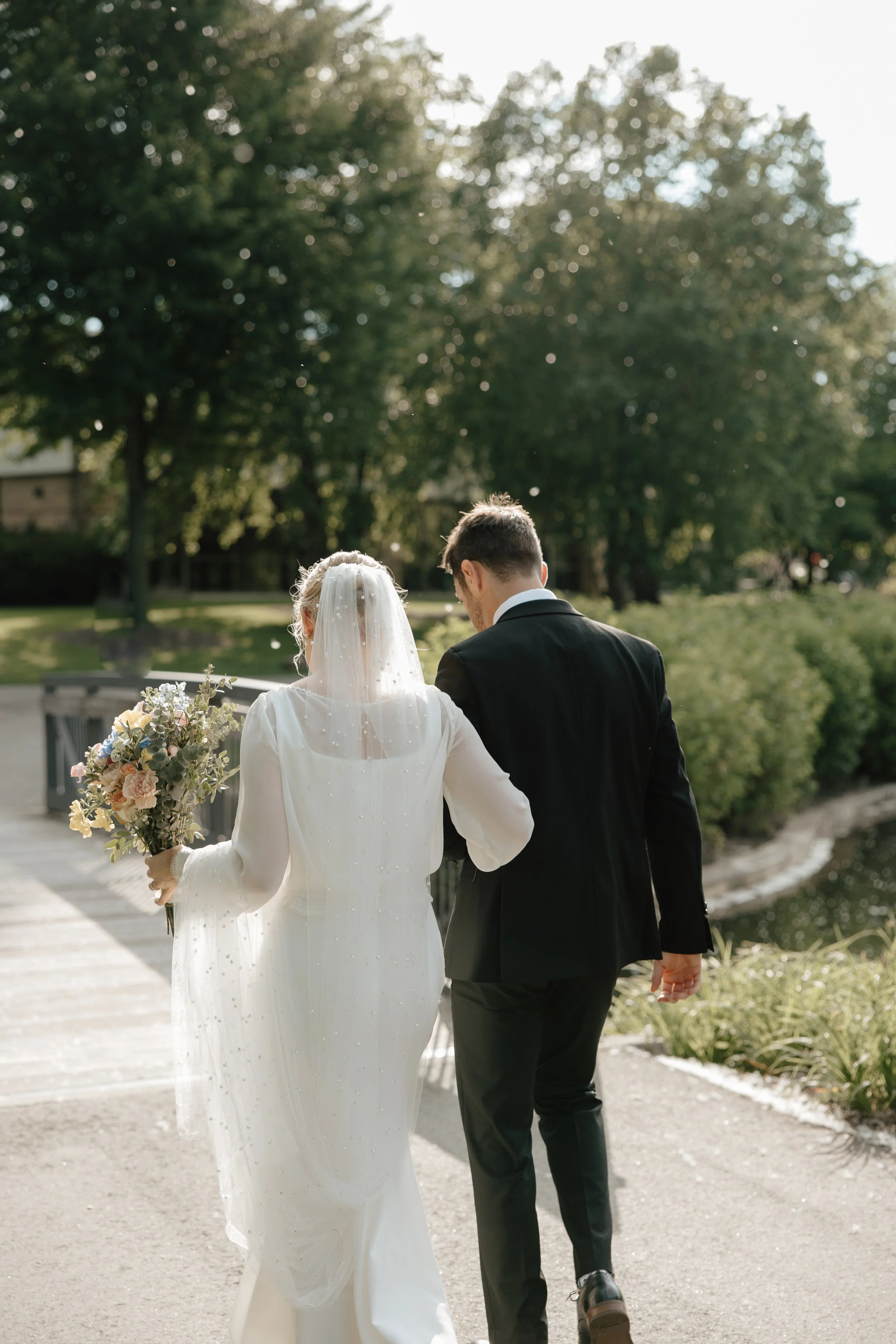 Bride and groom walking together by a river on their wedding day, with the bride holding a bouquet of flowers.