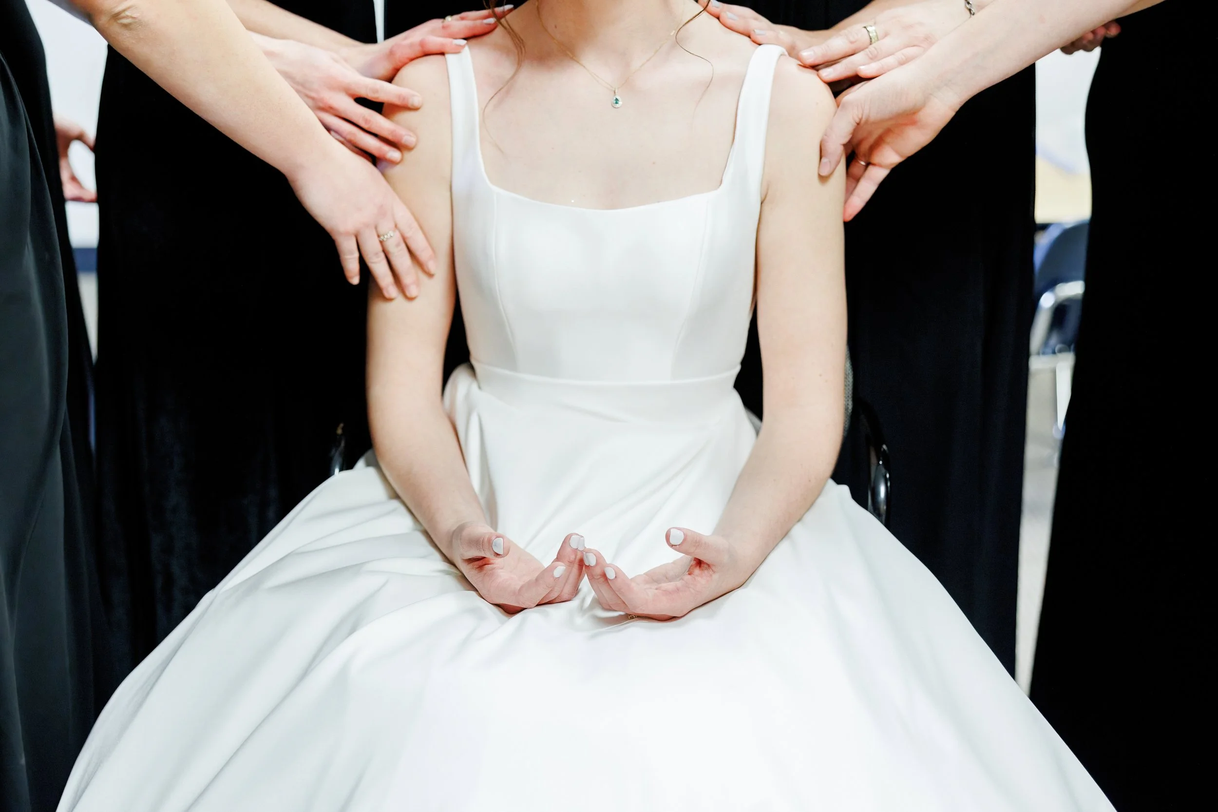 A bride sits with her hands in a meditation pose, surrounded by people touching her shoulders, wearing a white wedding dress and a necklace with a pendant.