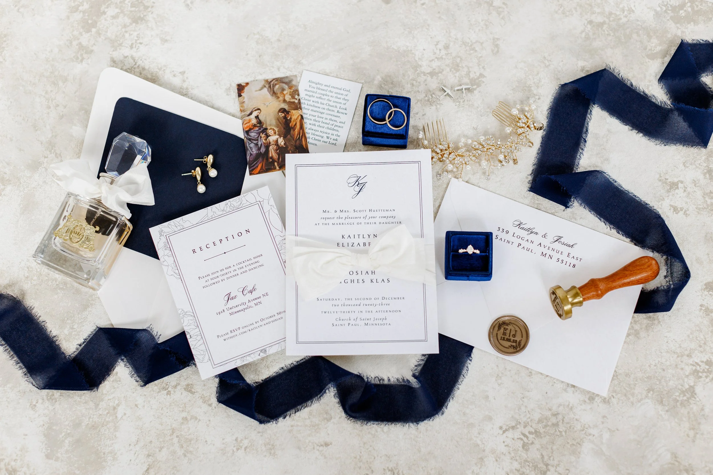 Wedding invitation, jewelry, perfume, and decorative items arranged on a light surface with a navy ribbon.