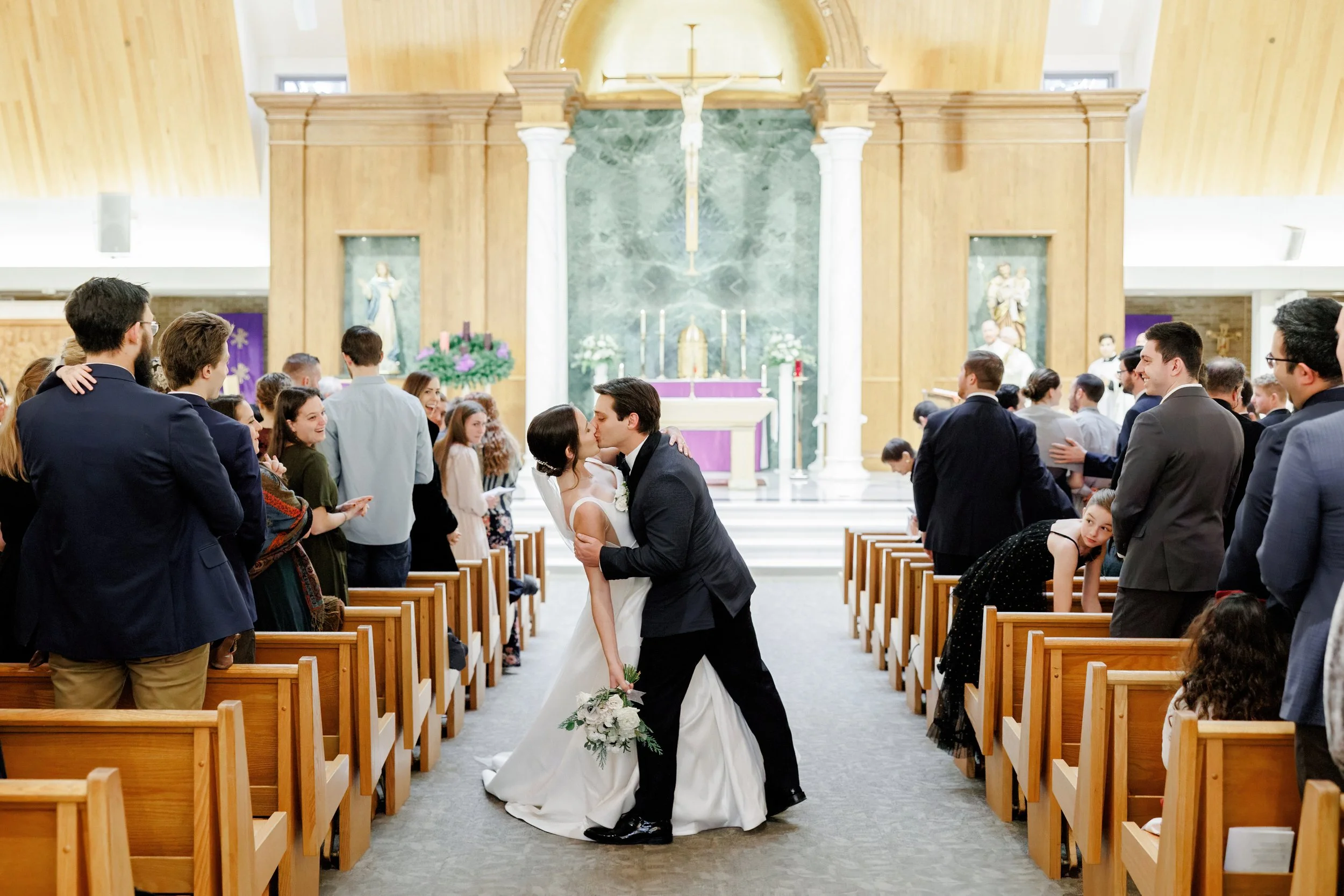 A newlywed couple sharing a kiss in a church during their wedding ceremony, surrounded by guests seated on wooden pews.