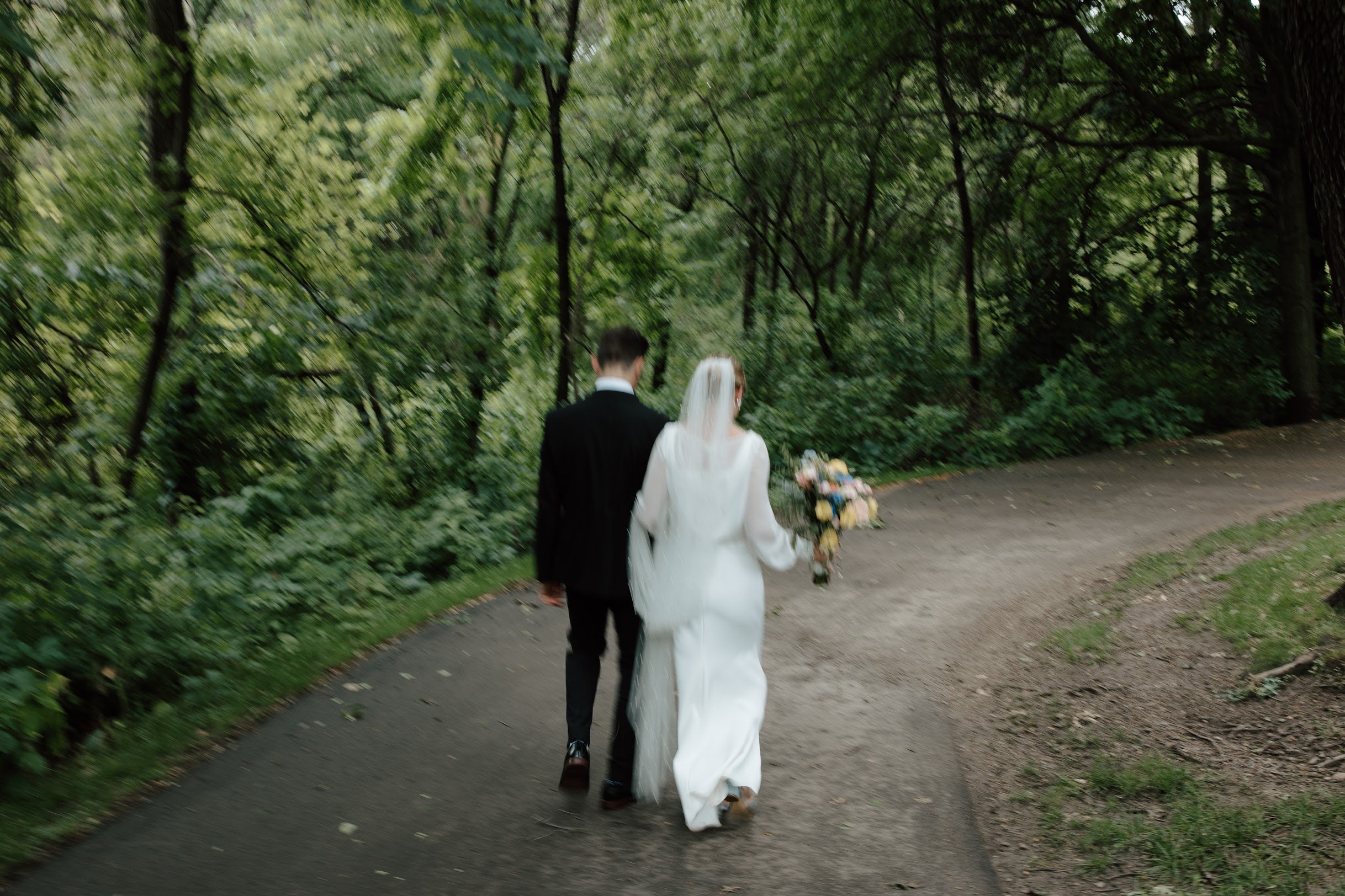 A bride in a white wedding dress and veil holding a bouquet walking with a groom in a black suit through a wooded park.