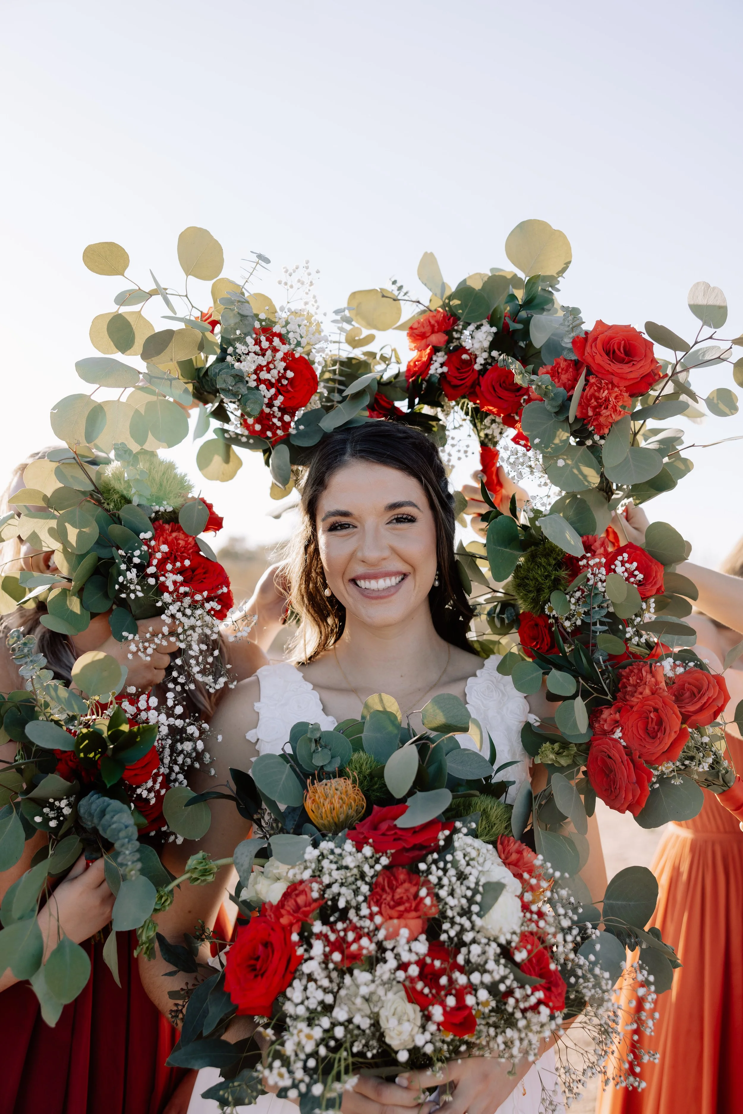 A woman smiling while holding a bouquet of red and white flowers, surrounded by a floral wreath with red roses, greenery, and baby's breath, with people behind her holding the wreath, outdoors during daytime.