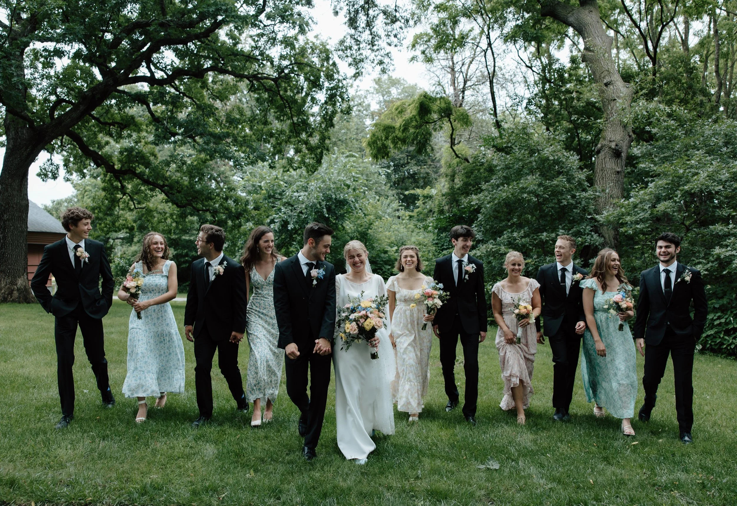 A group of people in wedding attire walking on a grassy field surrounded by trees, smiling and enjoying a celebration.