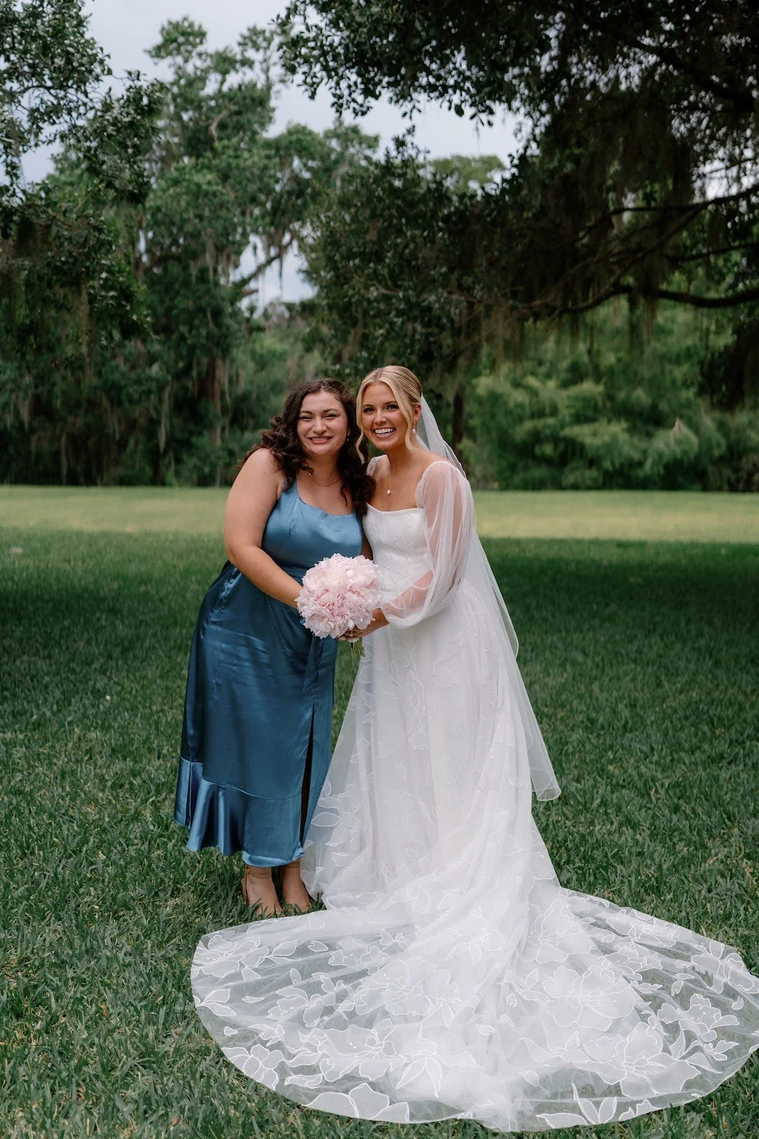 A bride in a white wedding dress and veil holding a bouquet of pink flowers, standing next to a woman in a blue dress, both smiling outdoors on a lush green lawn with trees in the background.