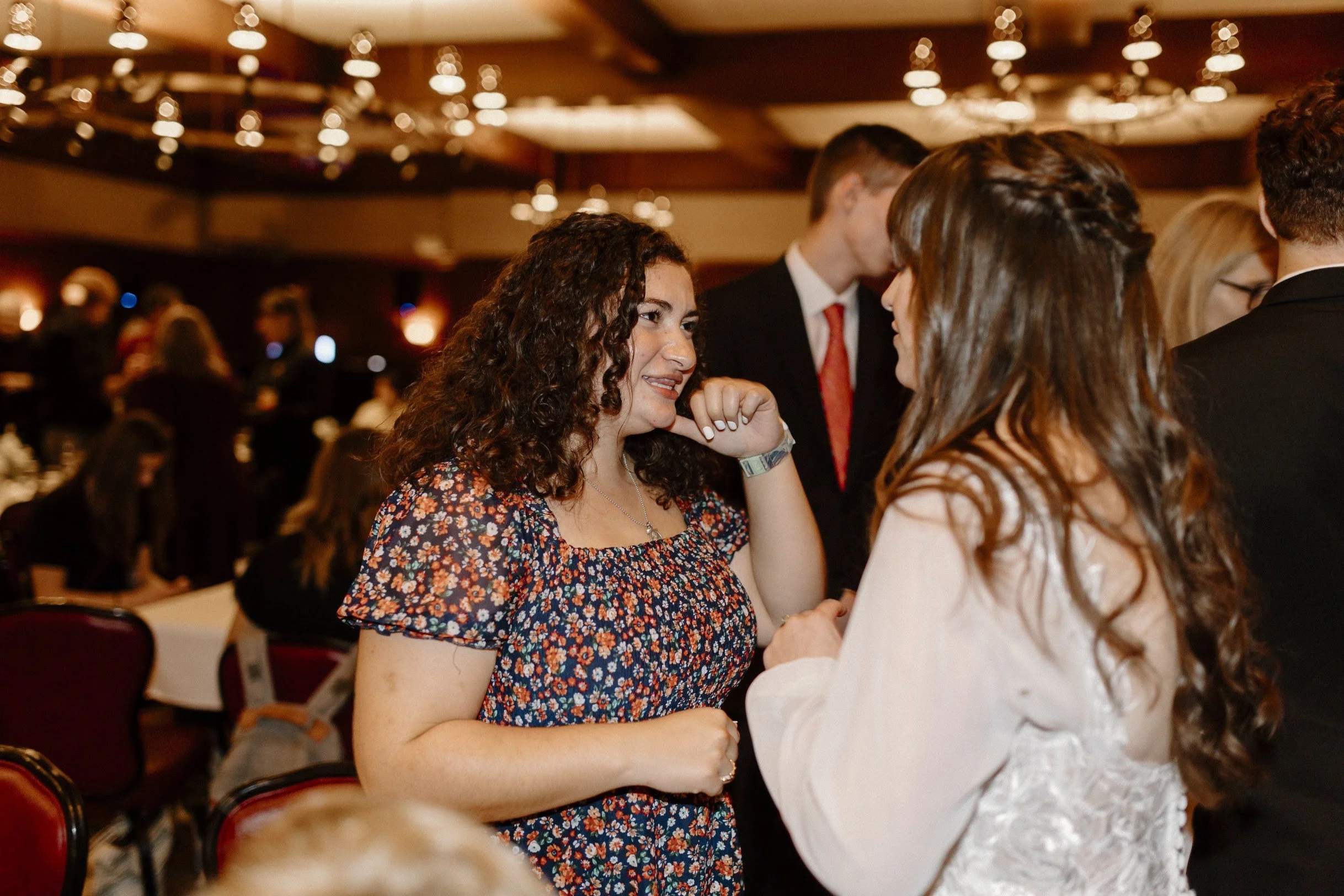 Two women with long brown and curly hair are talking and smiling at each other at a social event in a large, warmly lit banquet hall filled with people.