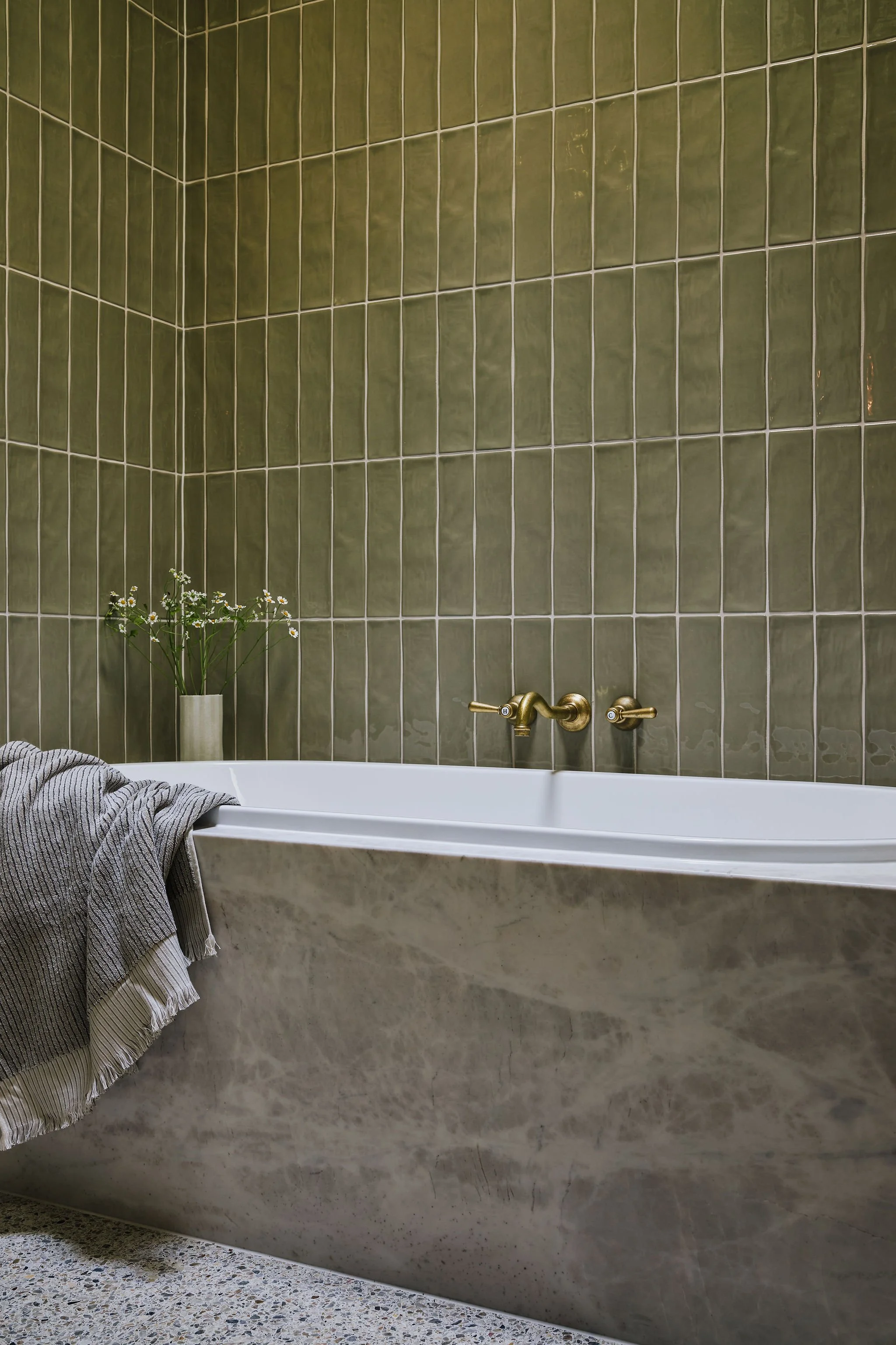 A bathroom with green tiled walls, a bathtub with a marble exterior, gold-colored faucet fixtures, a small white vase with white flowers, and a striped towel hanging over the edge of the bathtub.