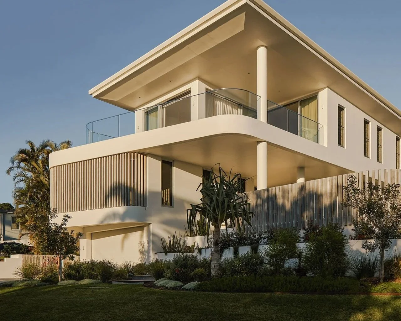 Modern two-story white house with large balconies, glass railings, and surrounding landscaped garden.