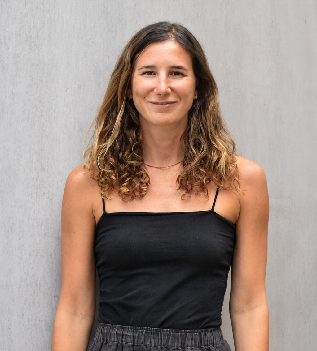 A woman with brown, curly hair wearing a black spaghetti strap top standing against a light gray textured wall.
