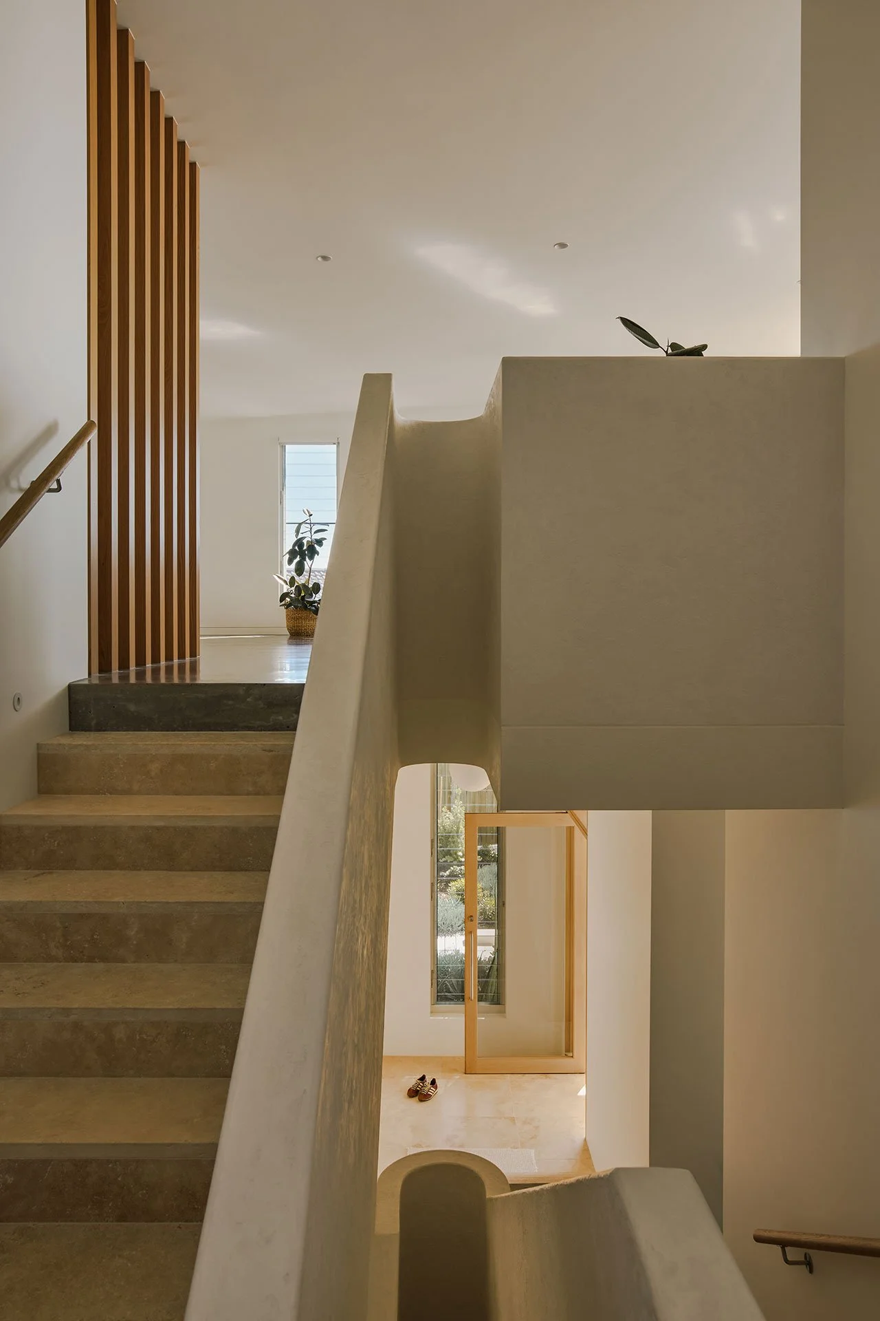 A staircase leading to a landing with a plants on the railing and a small window at the end of the hallway, with shoes on the floor near the door.