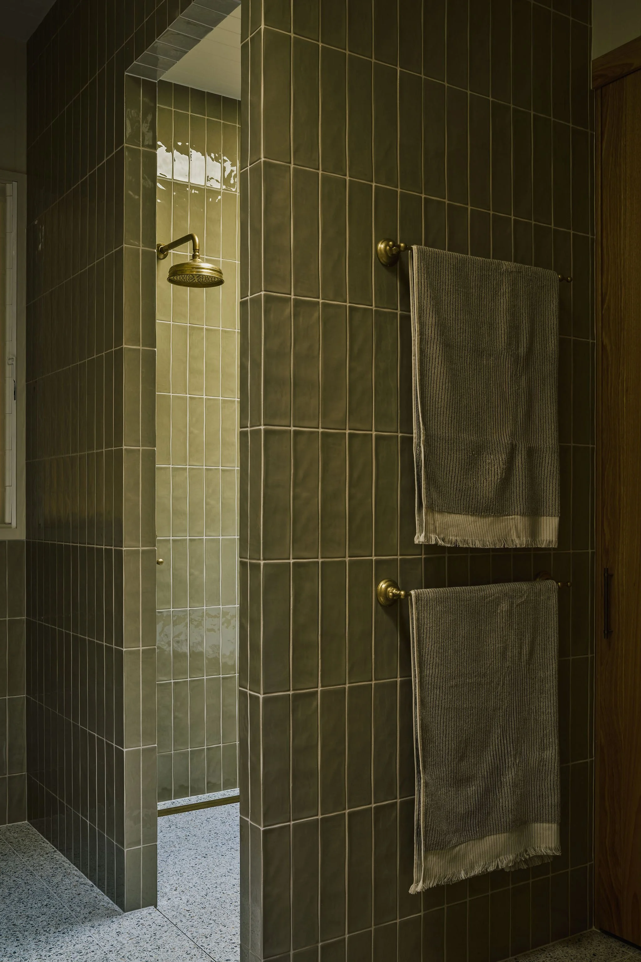 A bathroom shower with dark brown tiled walls, a bronze showerhead, and two matching dark towel racks with towels.