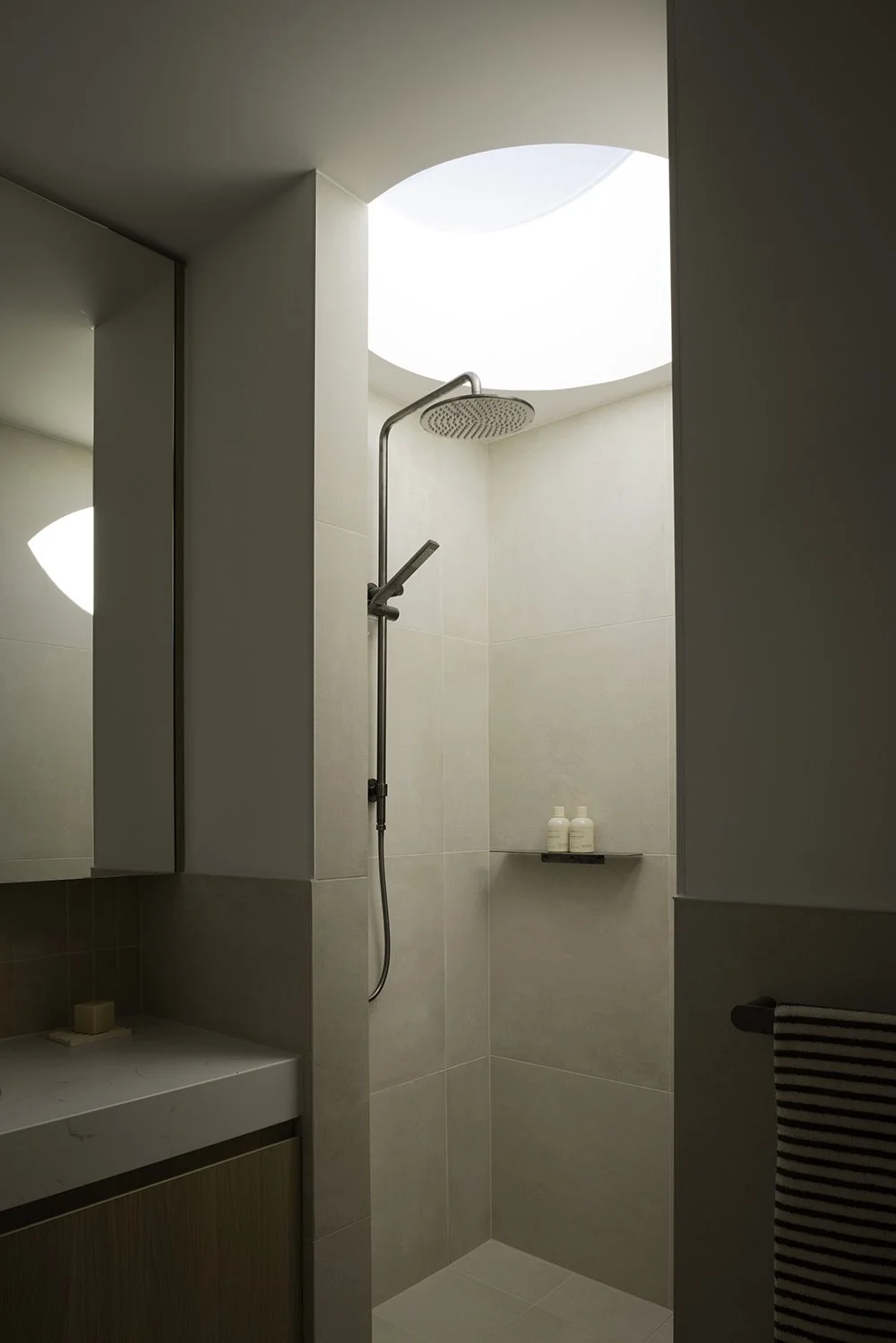 Modern minimalist shower area with a ceiling skylight, a wall-mounted showerhead, and a small shelf with toiletries, reflected in a mirror.