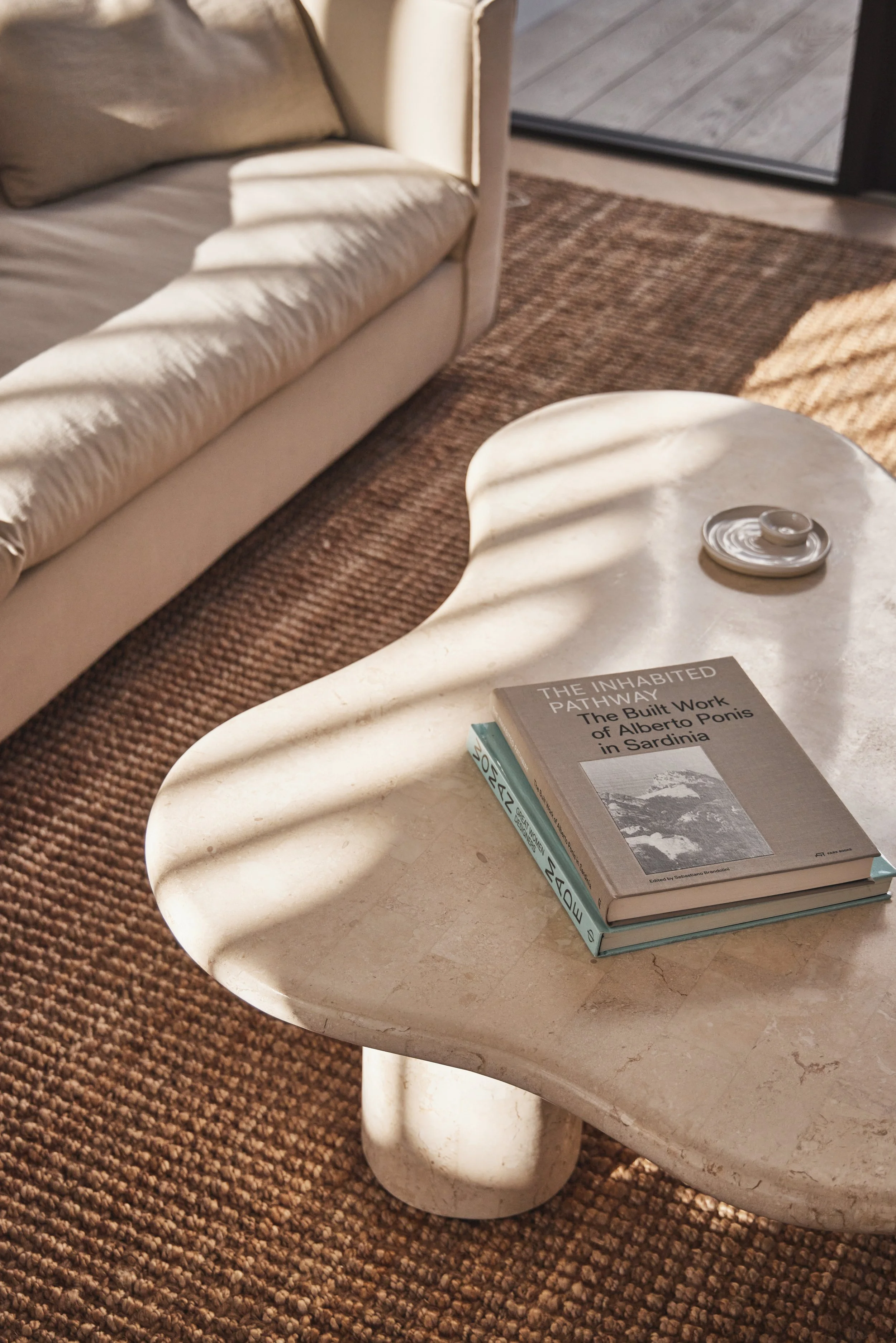 Close-up of a beige sofa, a white stone coffee table with rounded edges, a small silver dish, and two stacked books on the table. One book is titled 'The Inhabited Pathway'.