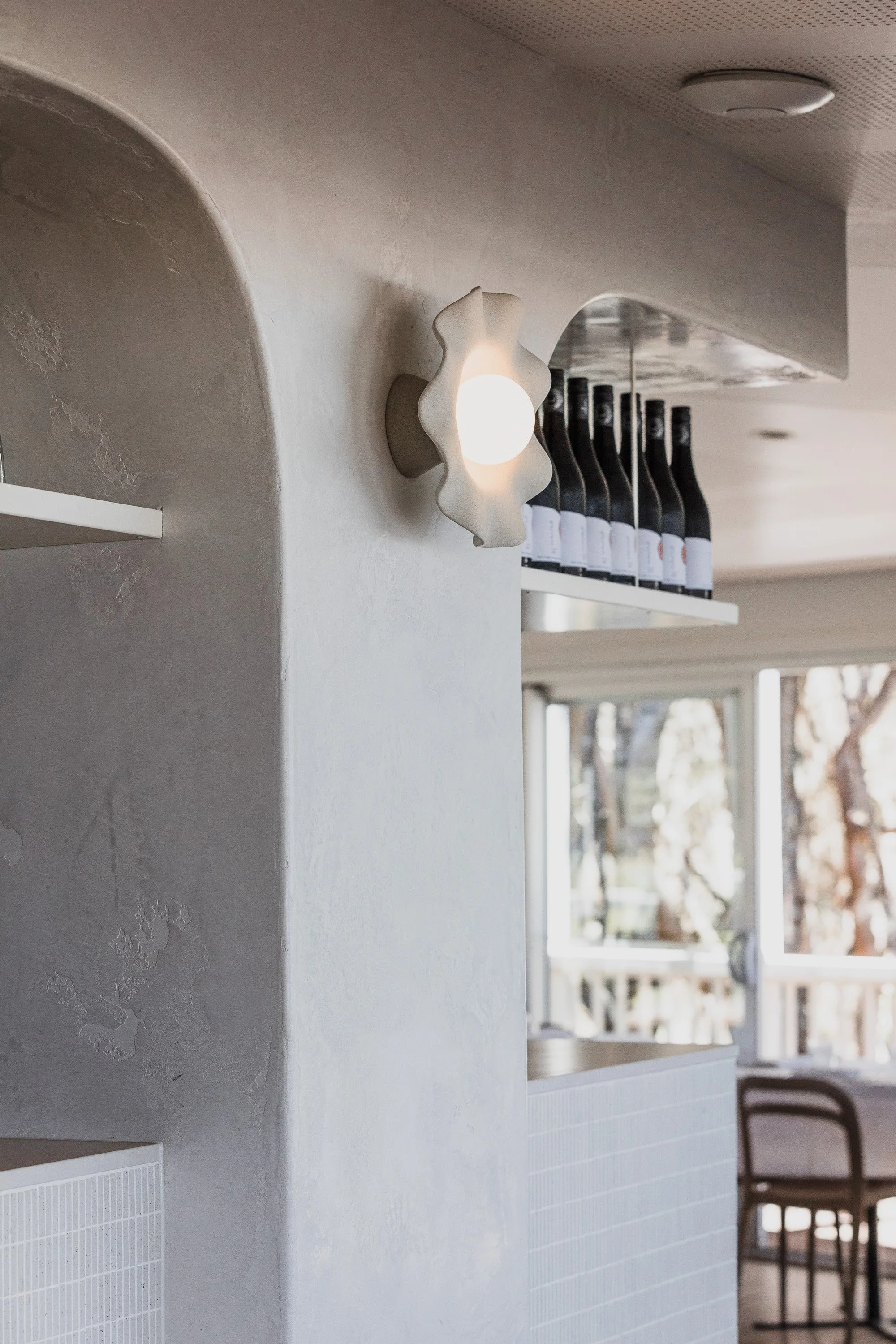 Interior view of a room with modern decor, including a white wall with textured paint, a decorative wall sconce with a white floral-shaped cover, a white shelf with bottles, a window with trees outside, and chairs around a table.