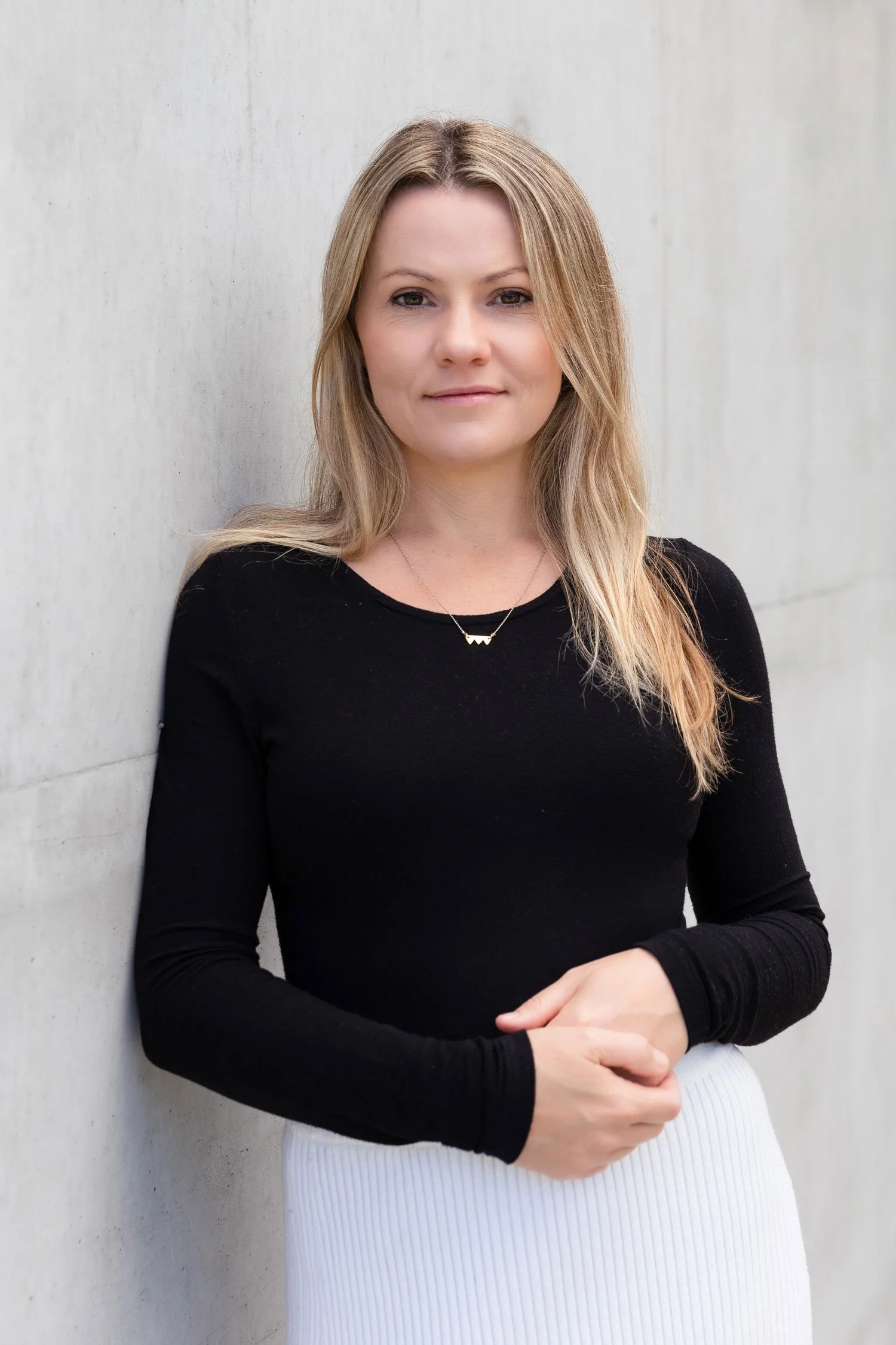 A woman with long blonde hair wearing a black top and a white skirt, standing against a light-colored wall.