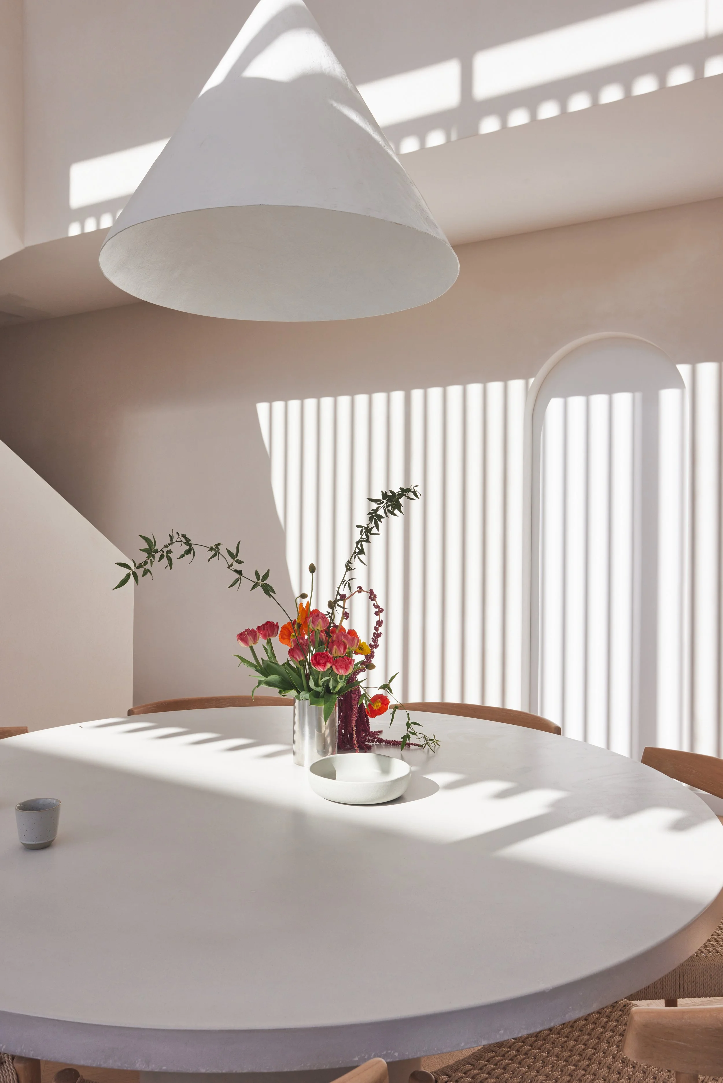 A white round dining table with a vase of colorful flowers and greenery in the center. Sunlight streams through blinds, casting shadows on the wall behind.