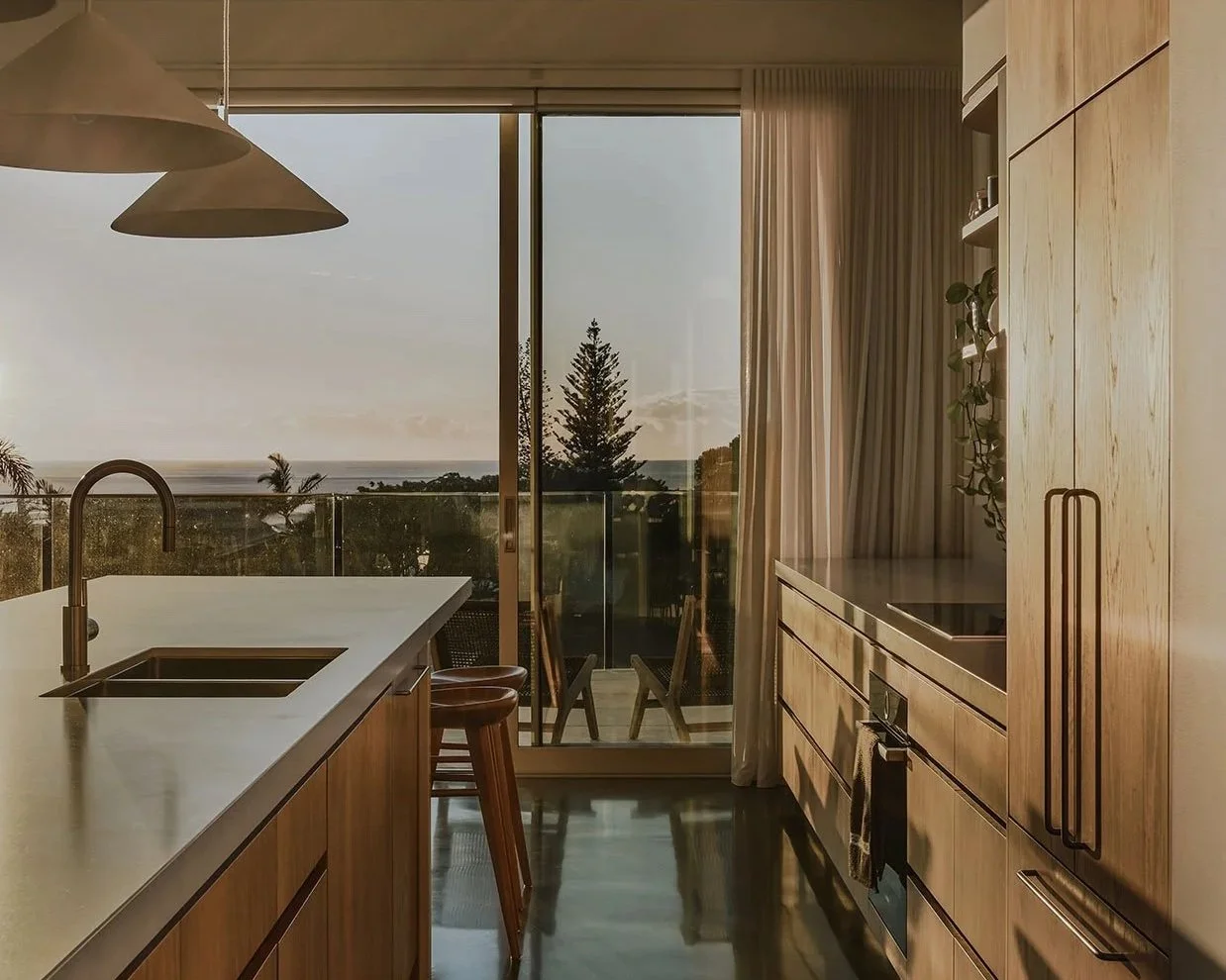 Modern kitchen with wooden cabinets, a white countertop, and a large sliding glass door leading to an outdoor balcony with a view of trees and the ocean at sunset.