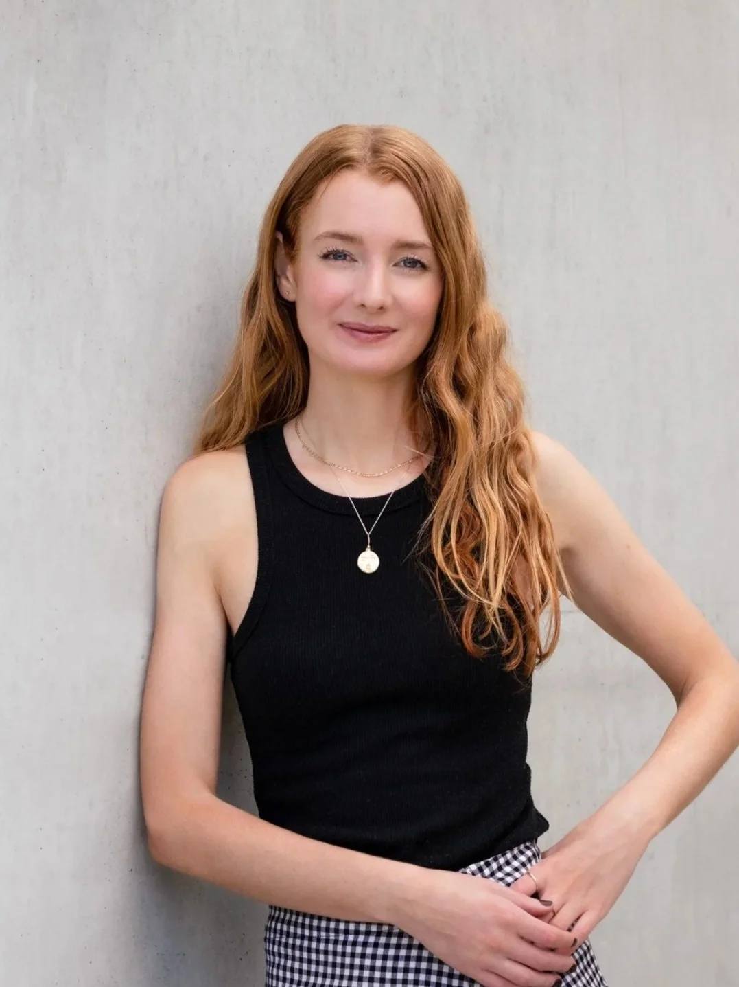A young woman with long wavy red hair, wearing a black sleeveless top and checkered pants, standing against a light gray plain wall, smiling softly.