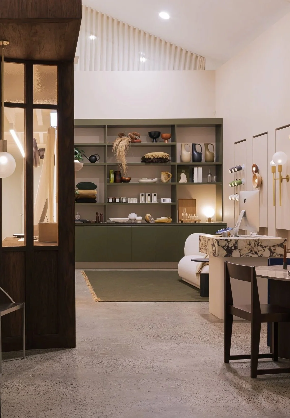 Interior of a modern store or showroom with shelves displaying ceramics, decor items, and accessories; a corner with a white sofa, a marble-topped desk, and a dark wooden dining chair; warm lighting and minimalist decor.