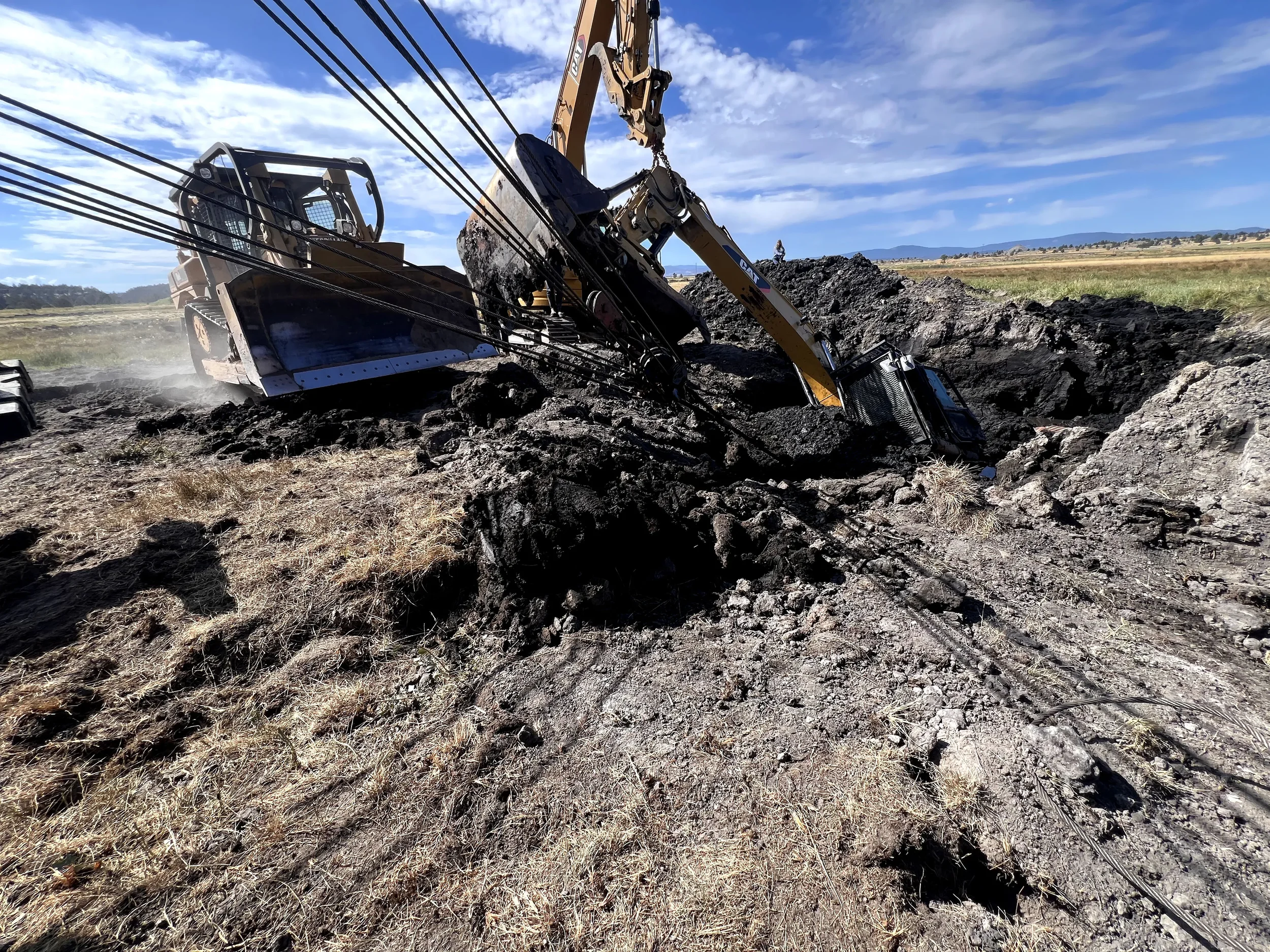 An excavator is partially submerged and tilted on its side in a large hole in a rural area with a grassy field and mountains in the background, under a blue sky with scattered clouds.