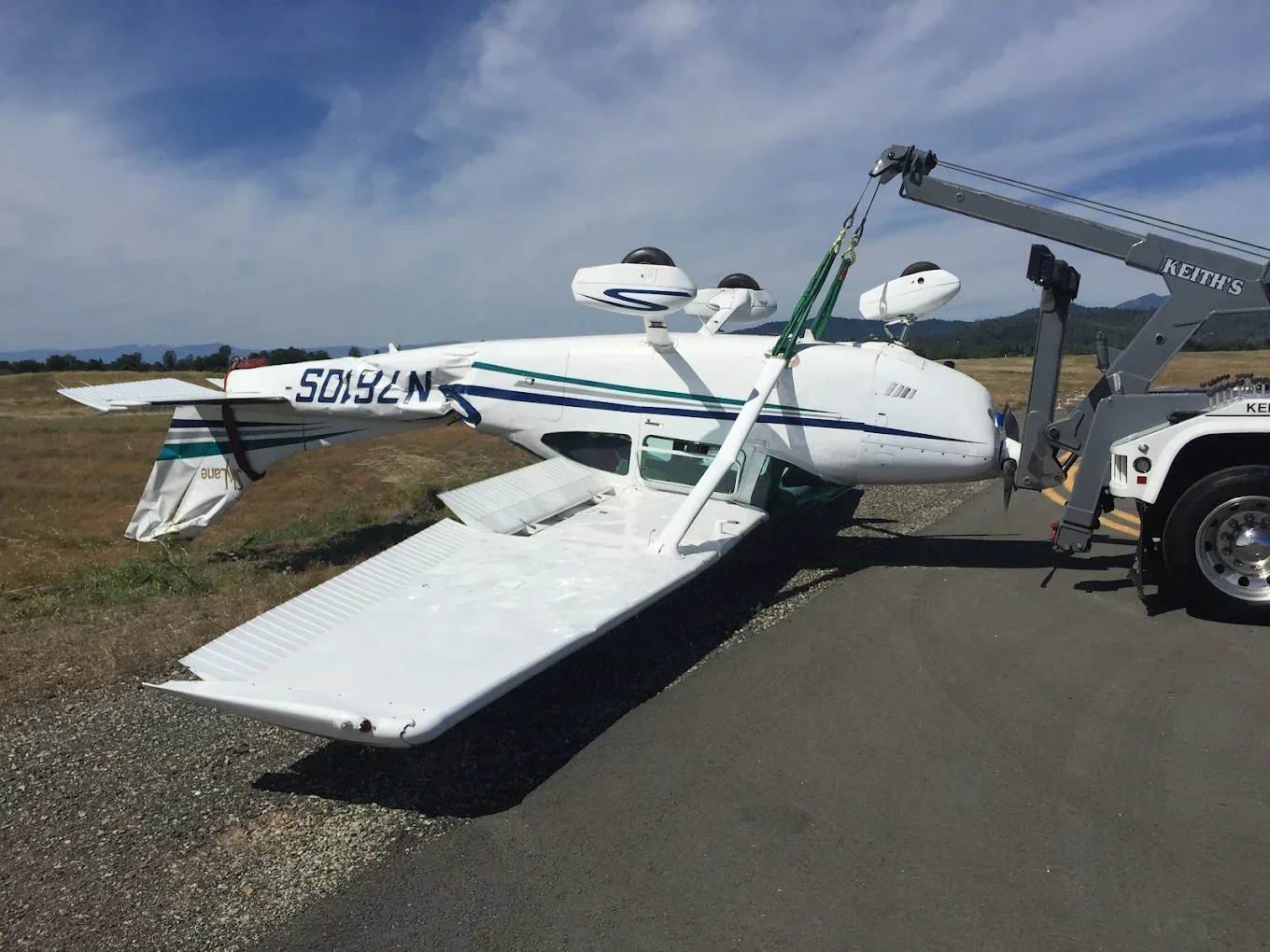 Small white airplane with blue and green stripes is damaged and partially collapsed on the roadside, being towed by a tow truck.