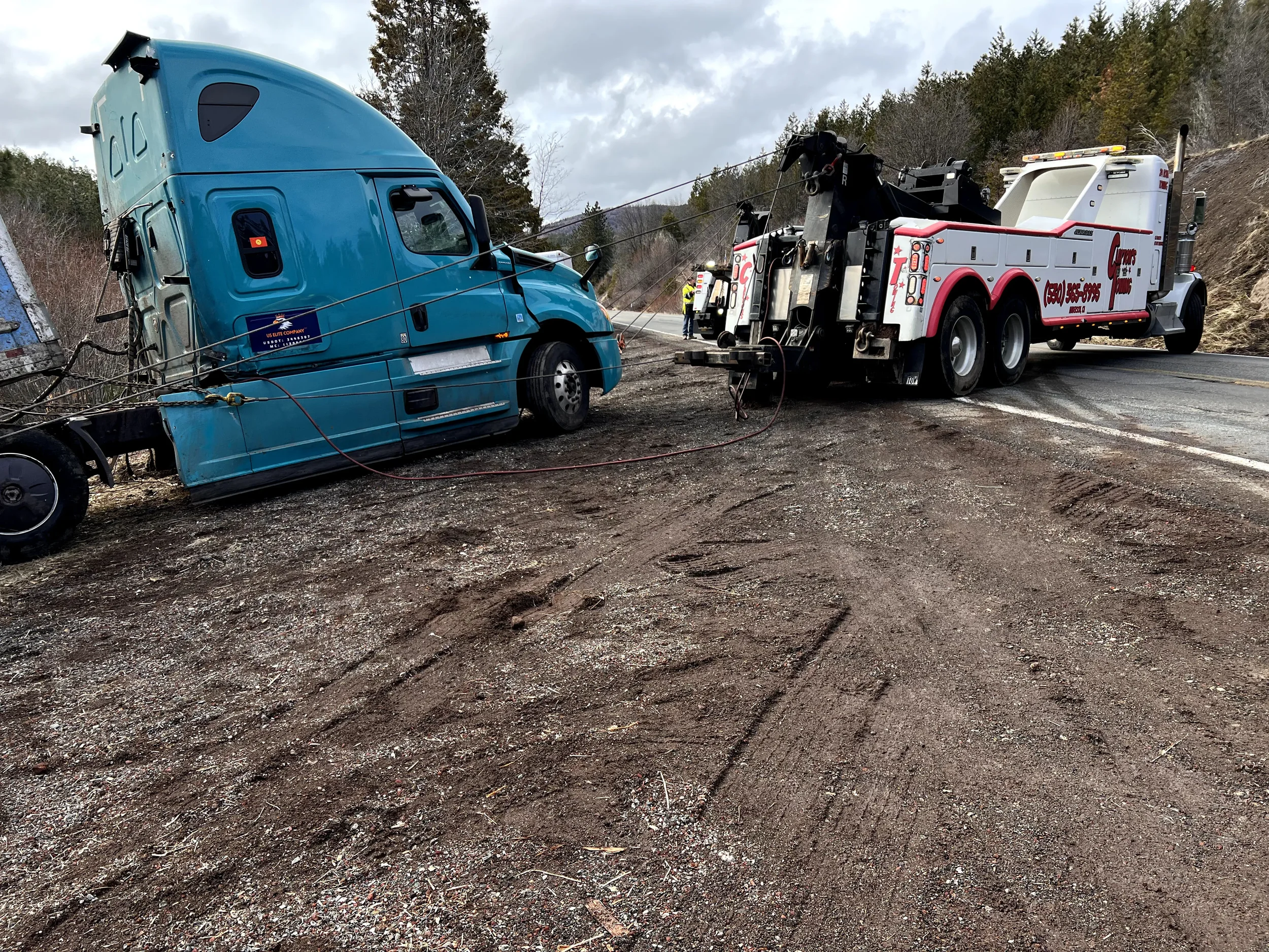 A semi-truck has crashed into a hillside and is being assisted by a tow truck on a rural road with cloudy skies.