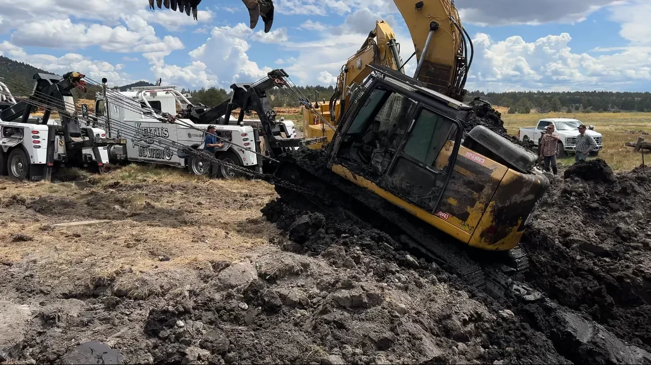 A yellow excavator has tipped over on uneven ground. Several tow trucks are attached to it using chains to tow it upright. Two people are standing nearby, observing the scene, with a field and a partly cloudy sky in the background.