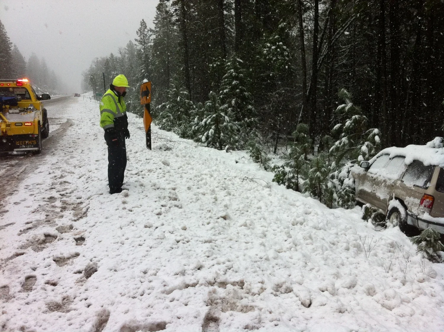 A person in a yellow safety jacket and helmet inspecting a snowy roadside where a car has slid off into the woods, with a snowplow vehicle nearby.