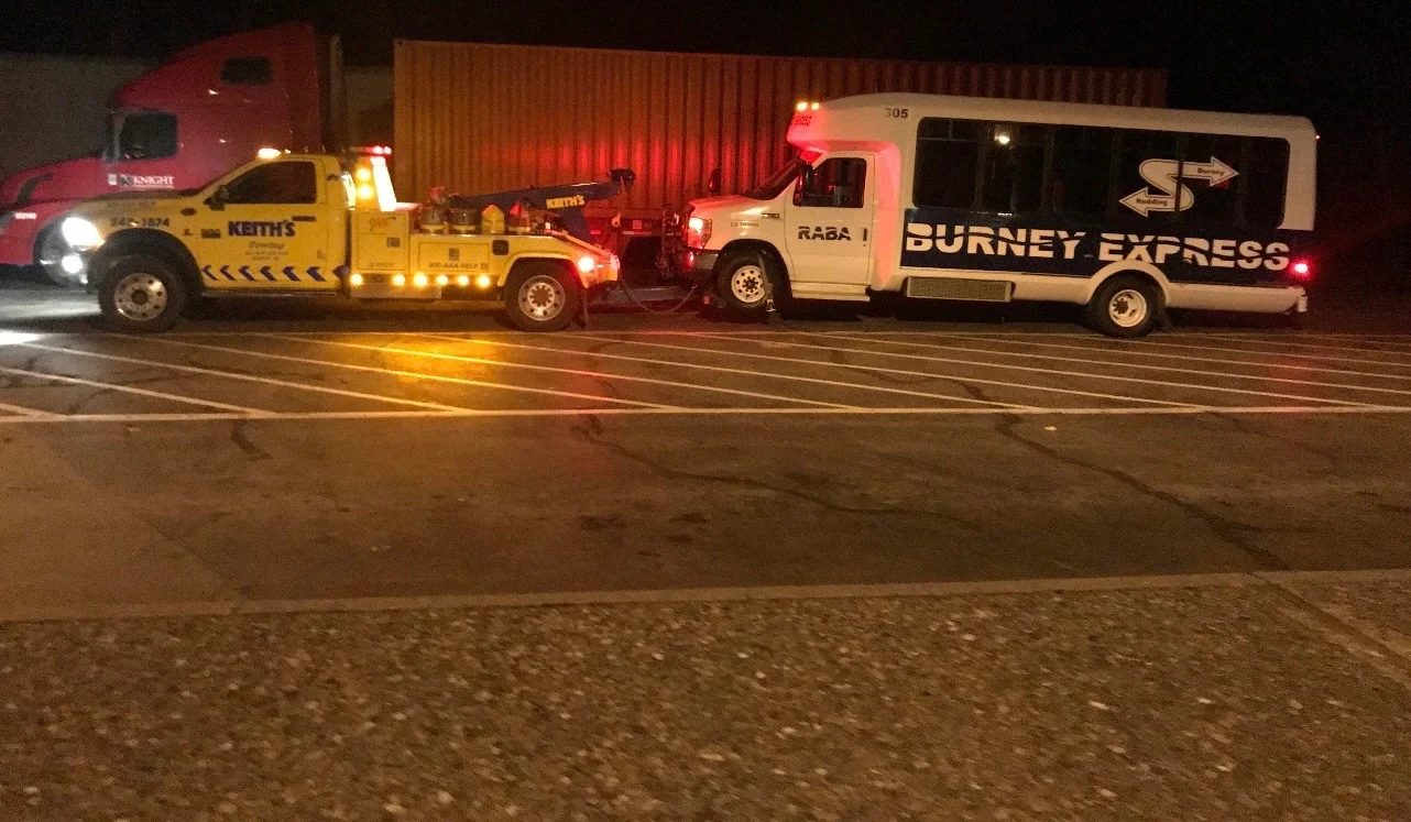A yellow tow truck attaching a white and black bus labeled 'Burney Express' to a tow cable in a parking lot at night.