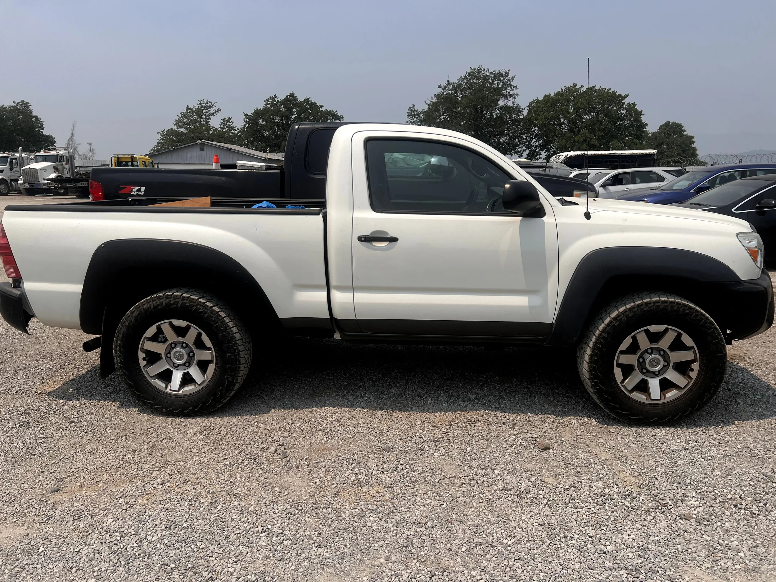 A white pickup truck parked on gravel with black wheel arches, black side mirror, and black front bumper. Multiple vehicles are visible in the background, and the sky is overcast.