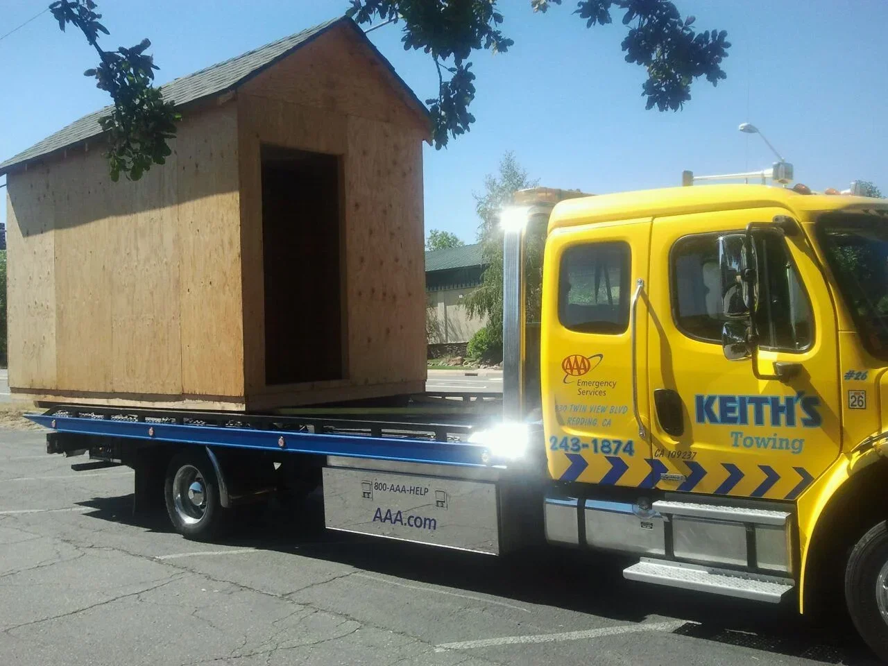 Yellow tow truck carrying a small wooden house on its flatbed in a parking lot.
