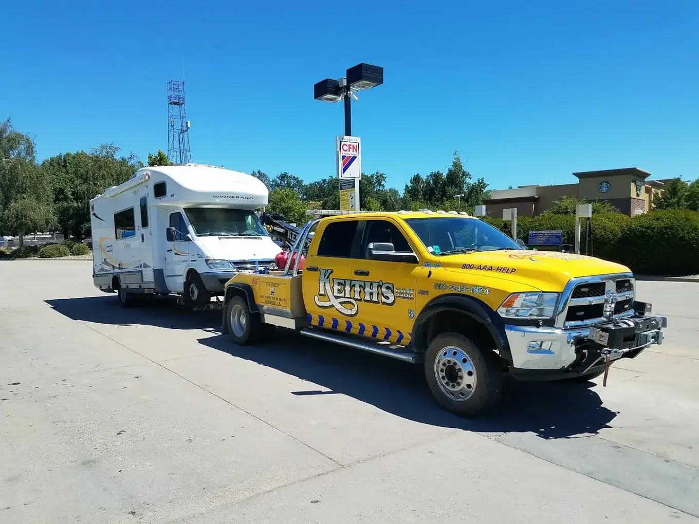 A yellow Keith's towing truck hauling a white Winnebago RV in a parking lot under a clear blue sky.