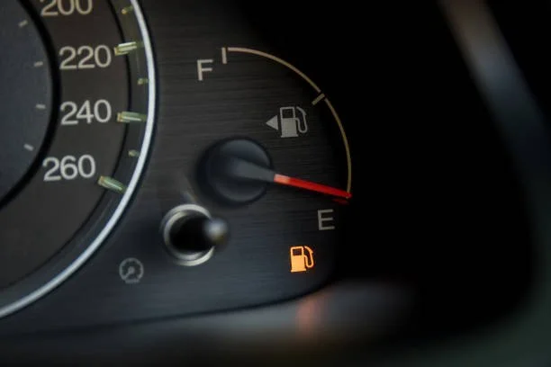 Car dashboard showing low fuel warning with fuel gauge pointing near empty and illuminated fuel warning light.