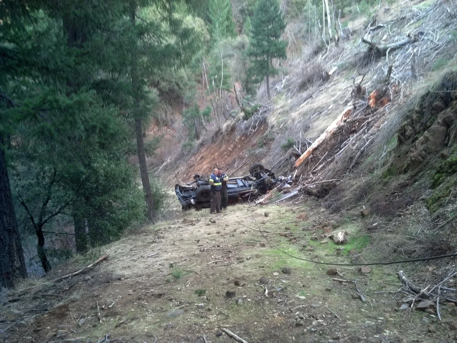Scene of a car crash on a wooded mountain trail, with an overturned vehicle against a steep slope and two people, possibly emergency responders or police, standing nearby.
